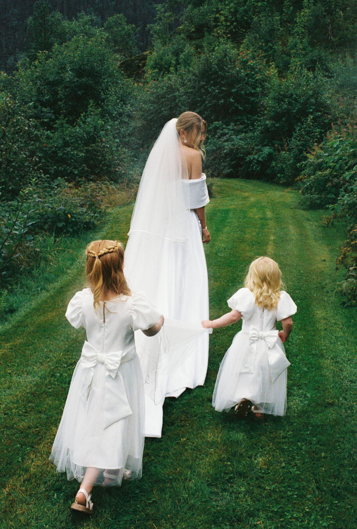 Bride in white gown and veil walks with two flower girls in matching white dresses on a lush green lawn.