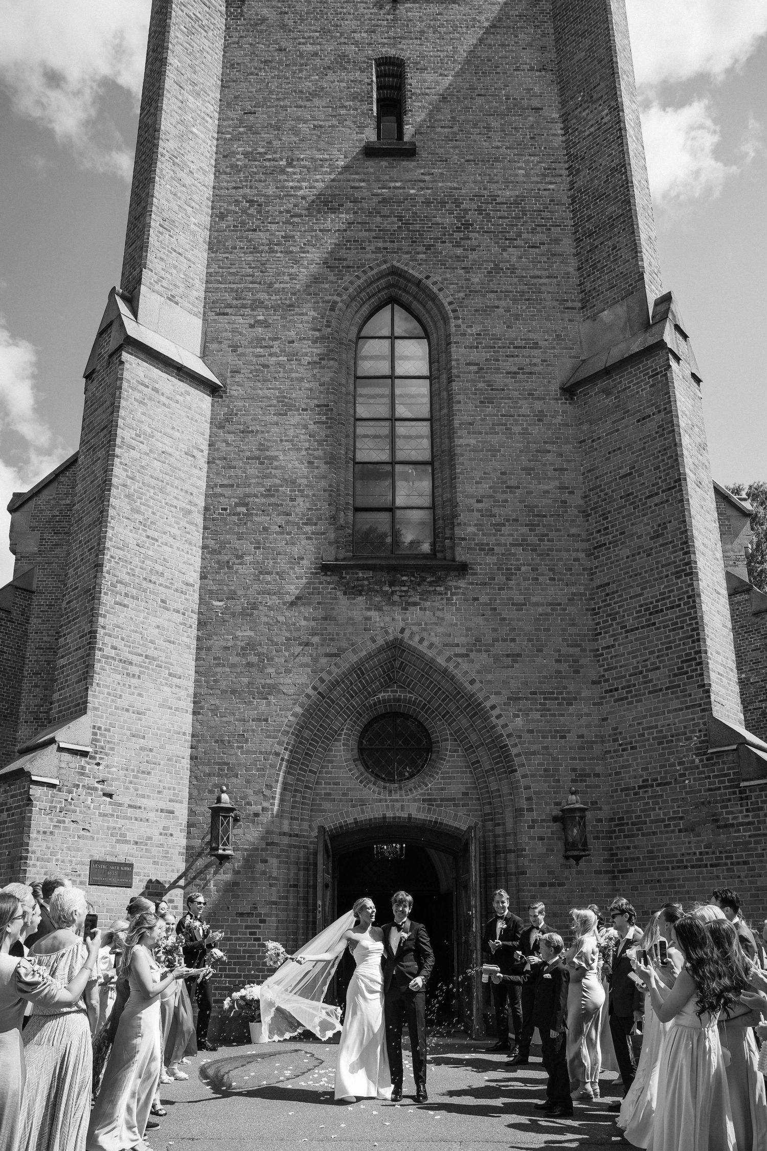 Black and white photo of a bride and groom exiting a gothic brick church surrounded by wedding guests.