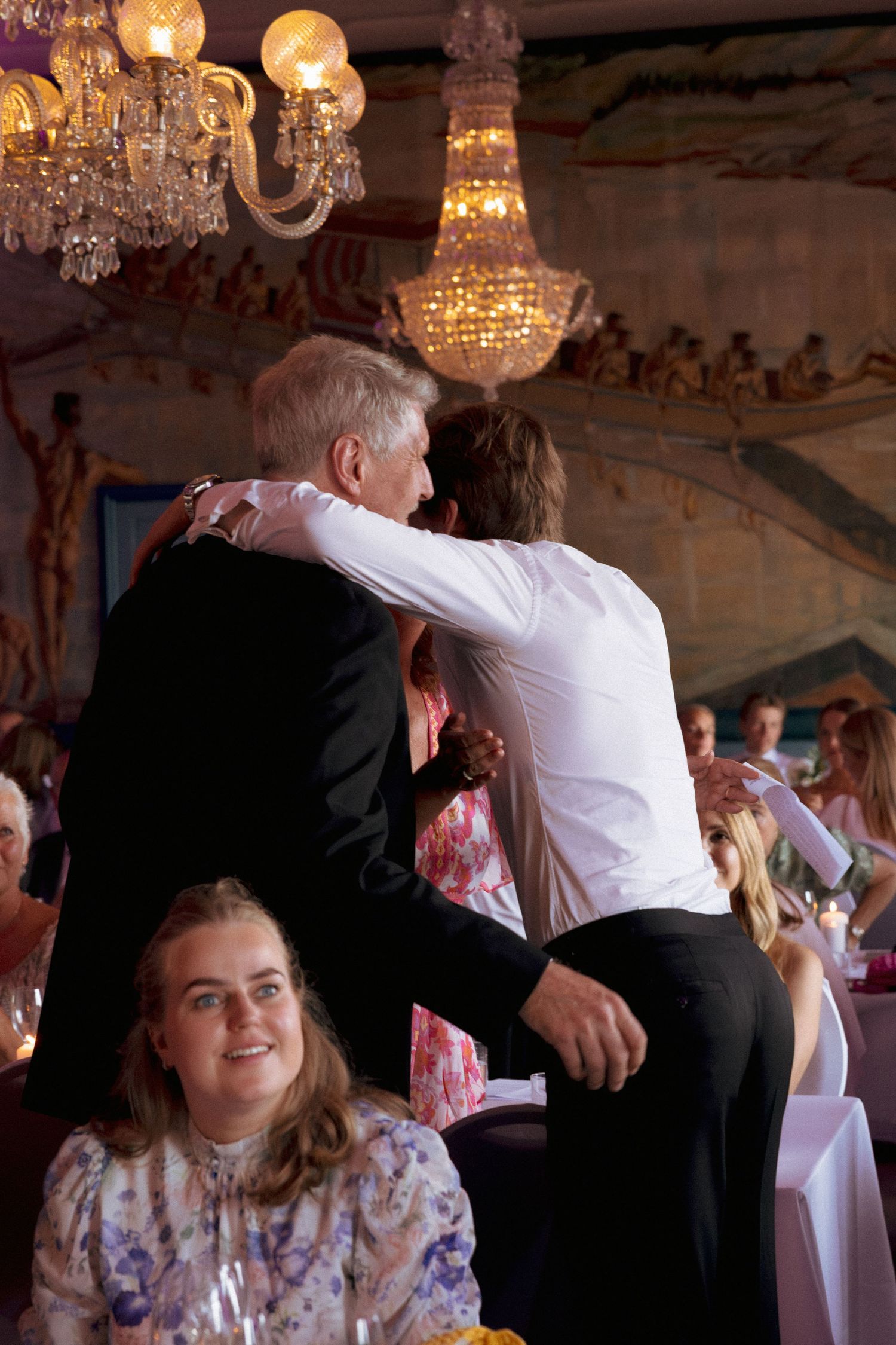 Guests embrace and celebrate at elegant wedding reception under crystal chandeliers in ornate venue.