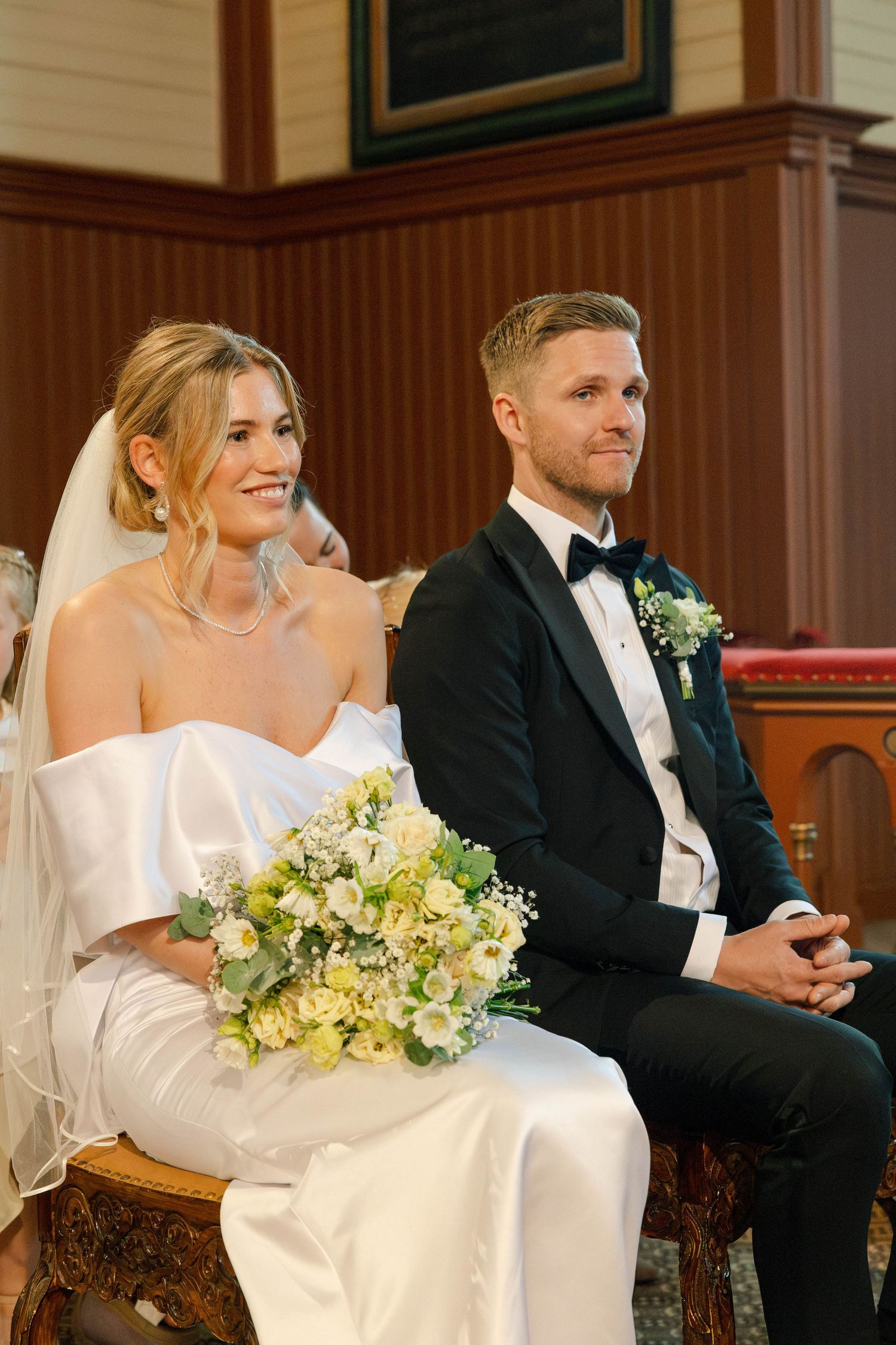 Smiling bride holding a white and yellow bouquet sits beside her groom in black tuxedo during wedding ceremony.