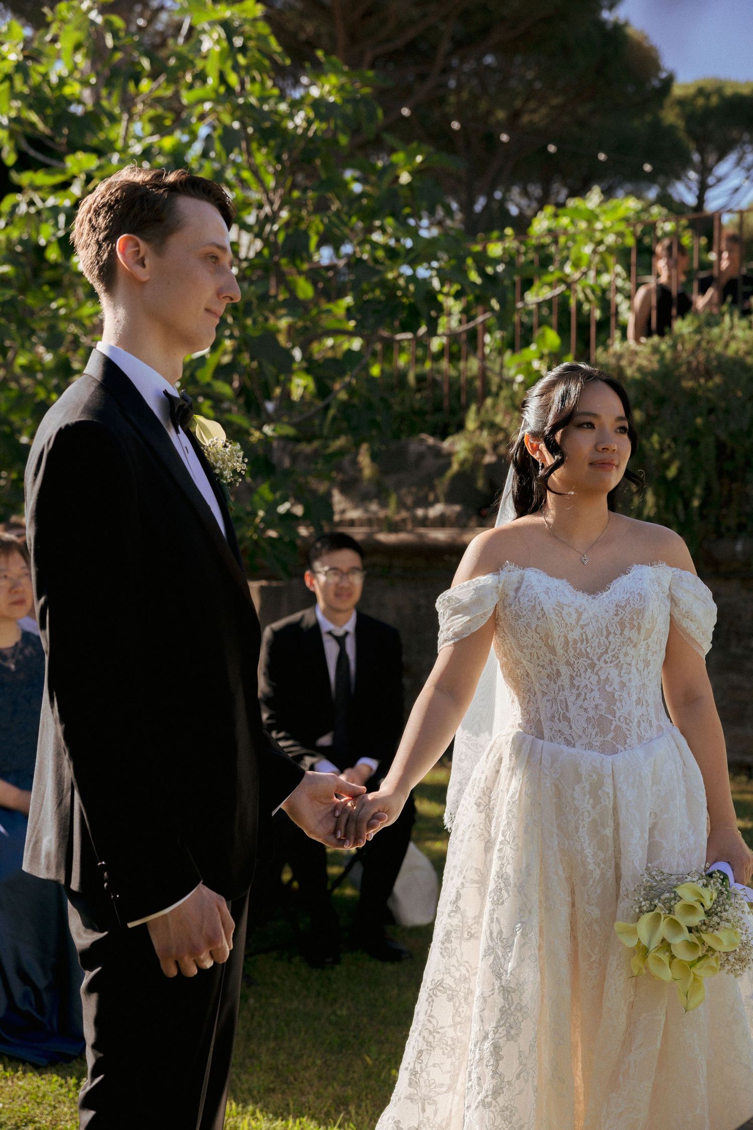 Bride and groom hold hands during outdoor wedding ceremony surrounded by lush greenery and seated guests.