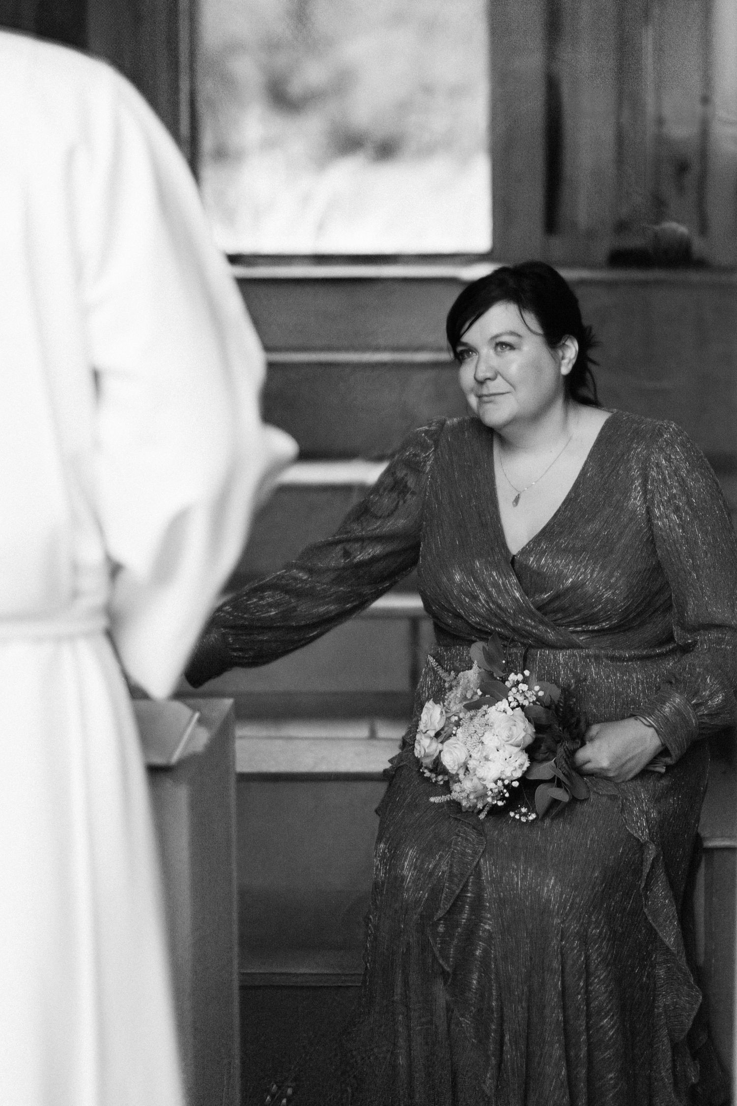 Black and white portrait of dark-haired bride holding white bouquet exchanging vows with officiant in rustic chapel.