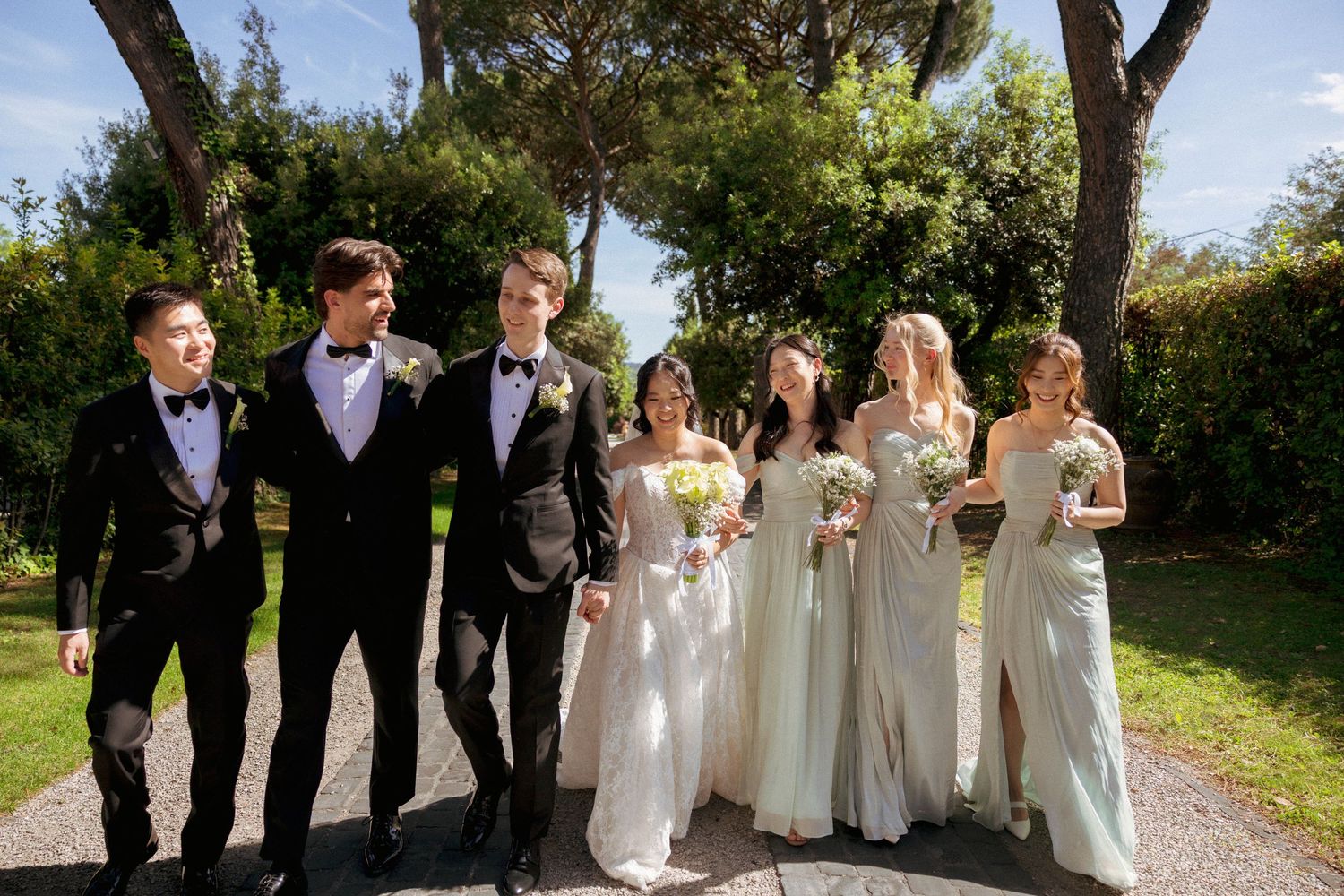 Wedding party walks outdoors, groomsmen in black tuxedos and bridesmaids in sage green gowns, with lush trees behind.