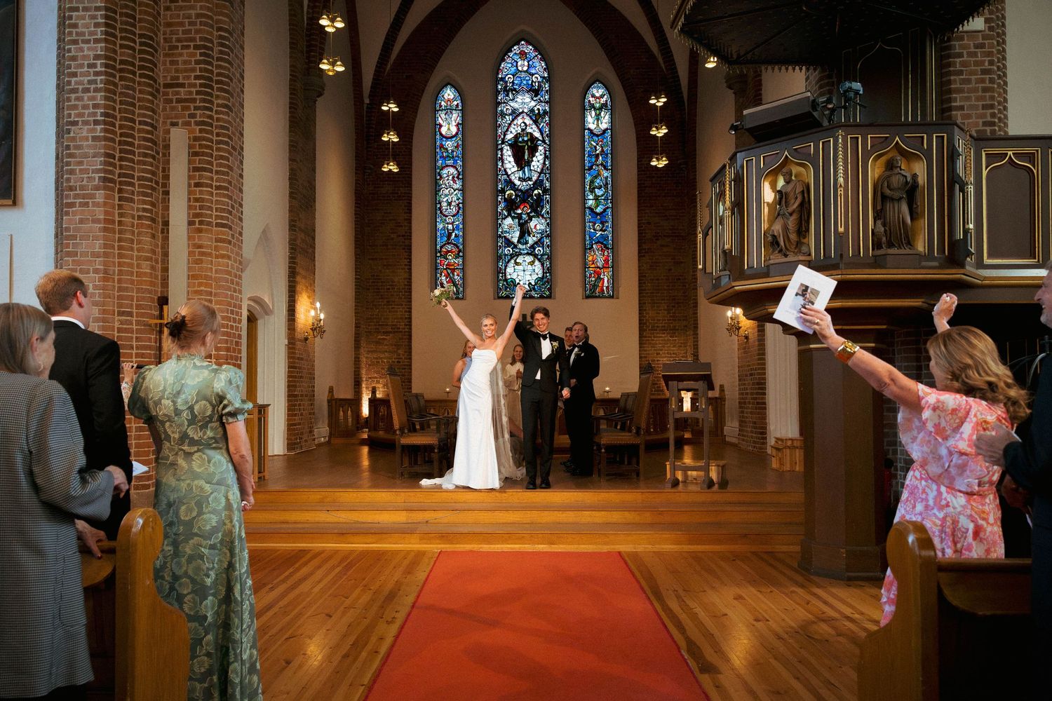 Joyful bride and groom celebrate at church altar with red carpet aisle and stunning stained glass windows.