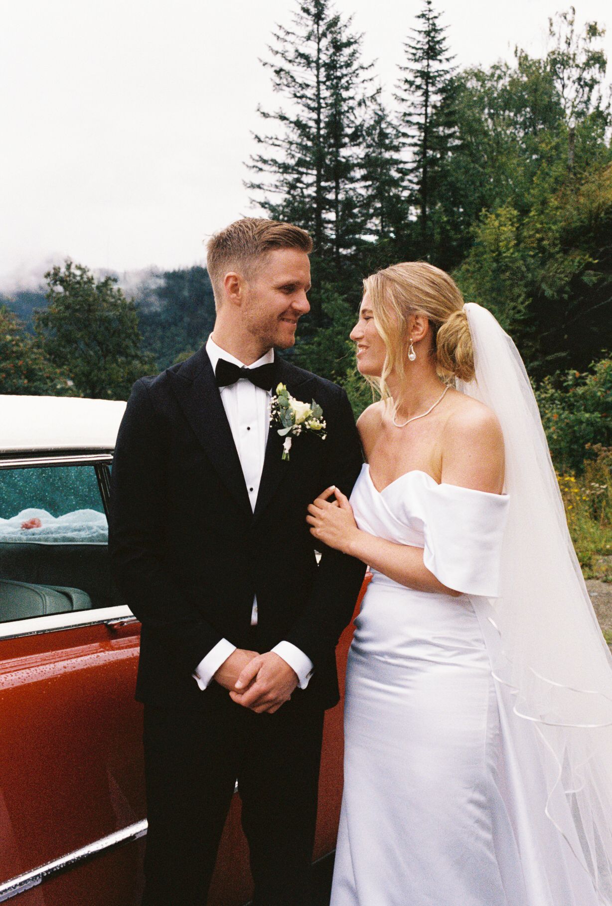 Smiling bride in off-shoulder white gown and groom in black tuxedo posing beside vintage red car outdoors.
