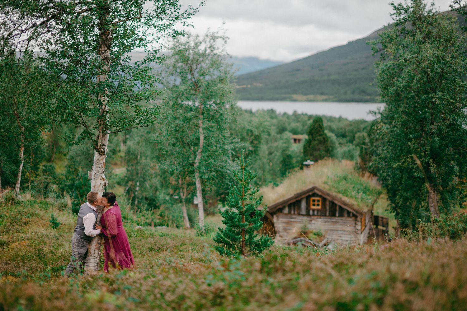 Couple embracing near birch trees with a grass-roofed cabin and mountain lake in a lush Norwegian landscape.