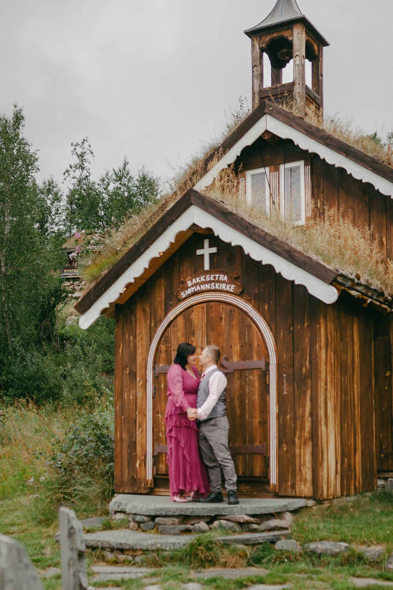 Couple sharing a kiss in front of a rustic wooden Norwegian chapel with a grass roof and cross.