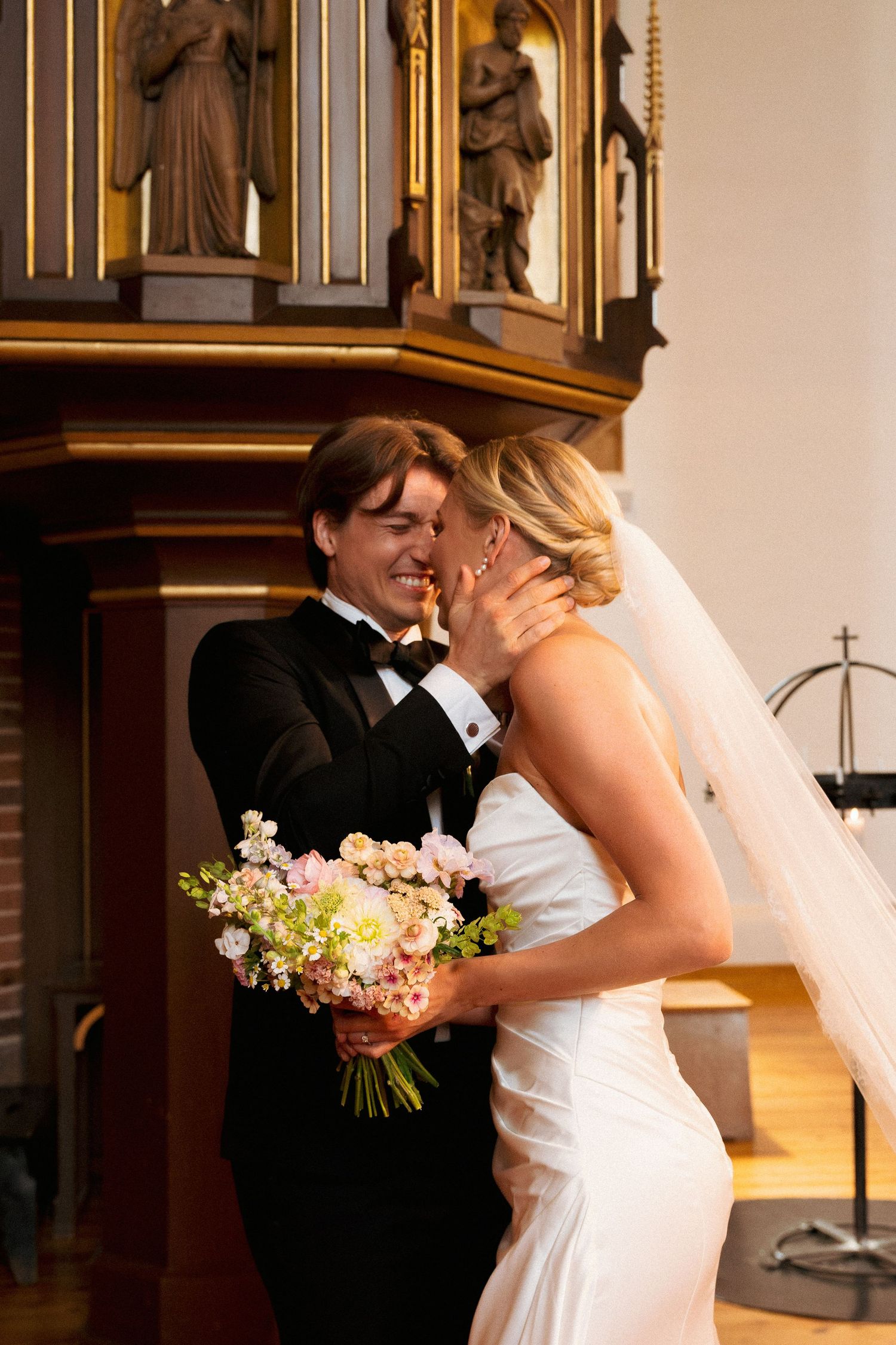 Smiling groom in tuxedo tenderly holds brides face during romantic church wedding ceremony, bride holds pastel bouquet.
