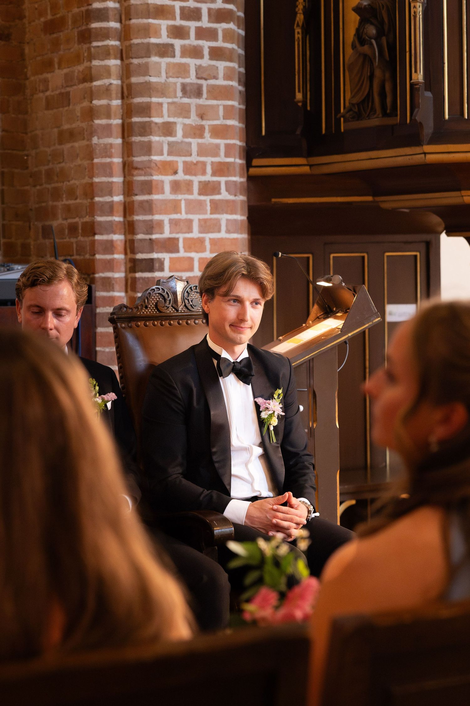 Groom in black tuxedo with bow tie sits smiling in ornate church chair during wedding ceremony.