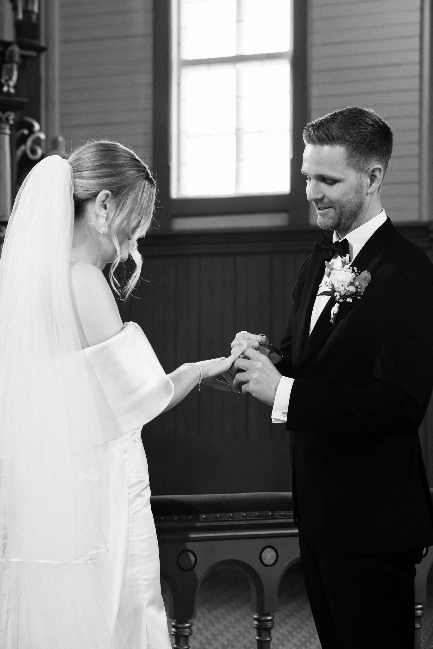 Bride placing wedding ring on grooms finger during an intimate black and white ceremony exchange.