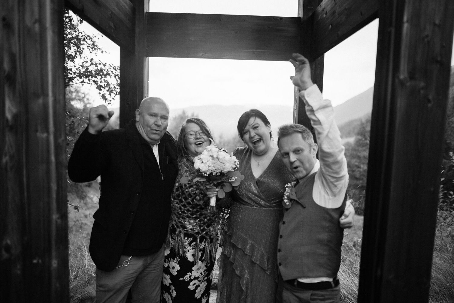 Black and white photo of joyful wedding party celebrating at rustic outdoor wooden structure.