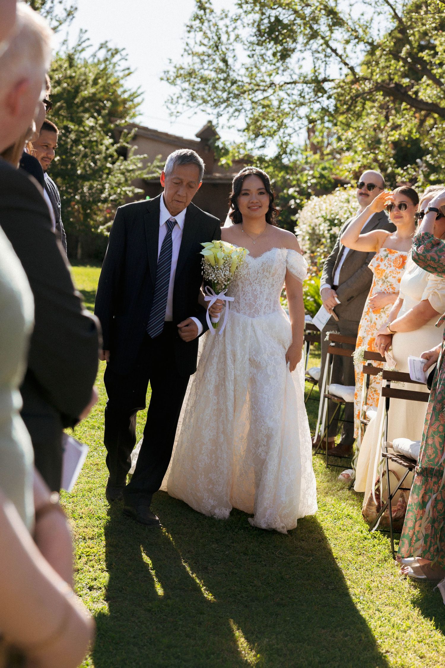 Bride in lace off-shoulder gown walks down outdoor aisle with escort on sunny wedding day.