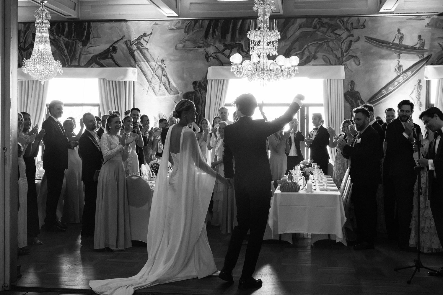 Black and white photo of bride and groom entering wedding reception as guests applaud in elegant venue.
