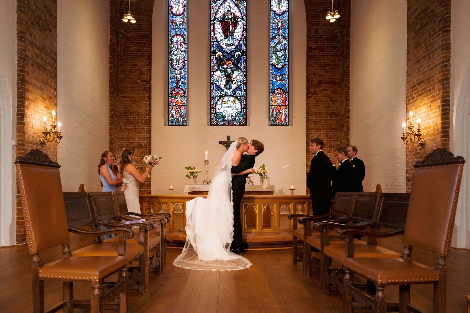 Bride and groom share first kiss at altar in elegant chapel with stained glass windows and brick walls.