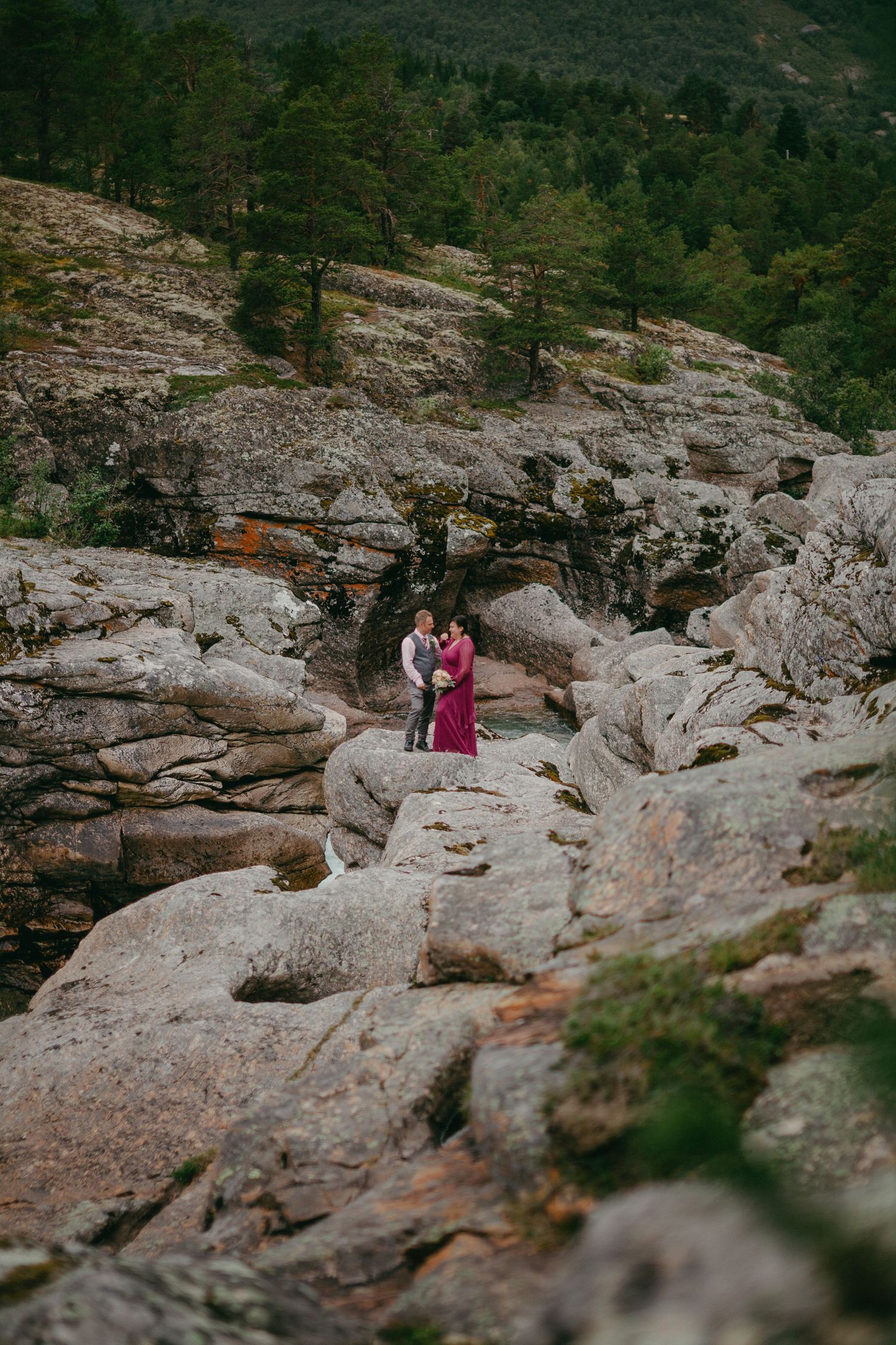 Wedding couple embracing on dramatic rocky landscape with pine forest backdrop during outdoor elopement ceremony.