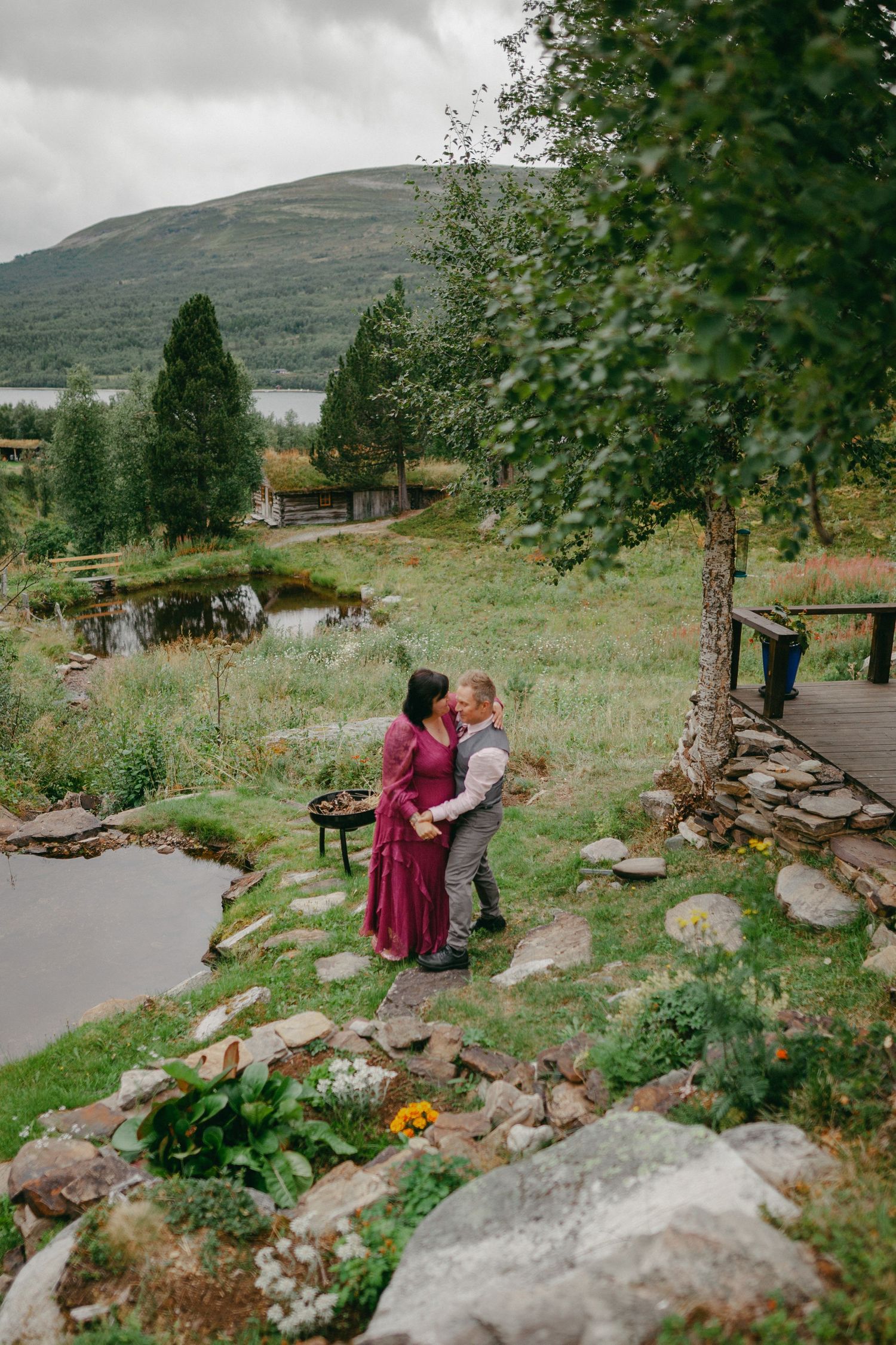 Couple embracing in a lush garden with a pond, green hills, and a lake backdrop on a cloudy day.