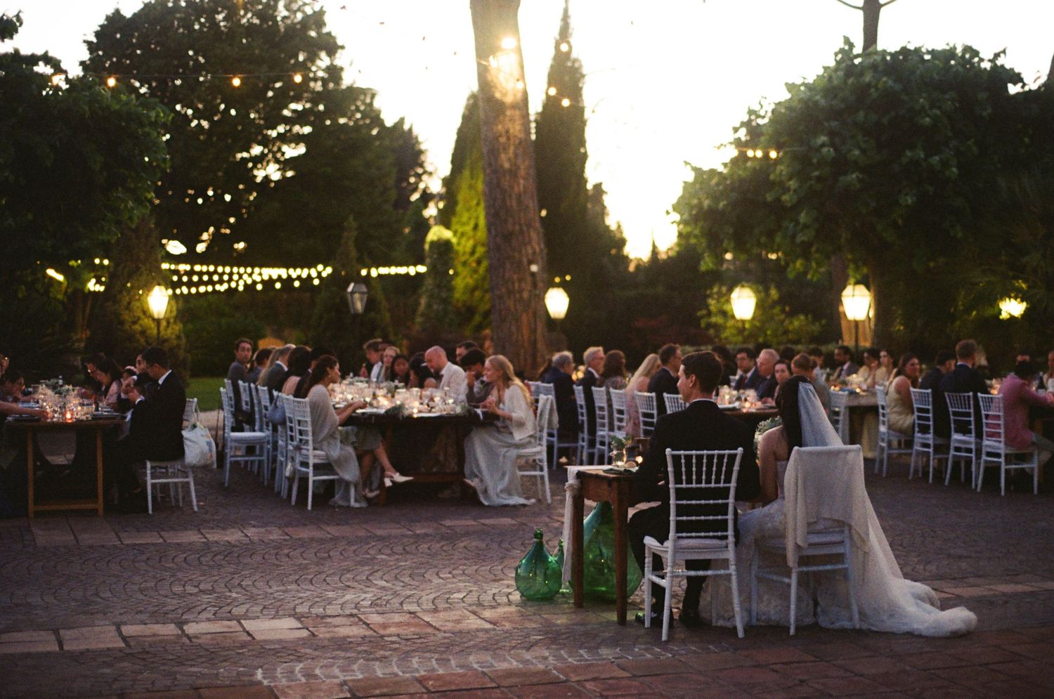 Elegant outdoor wedding reception at dusk with string lights and guests dining at long tables.