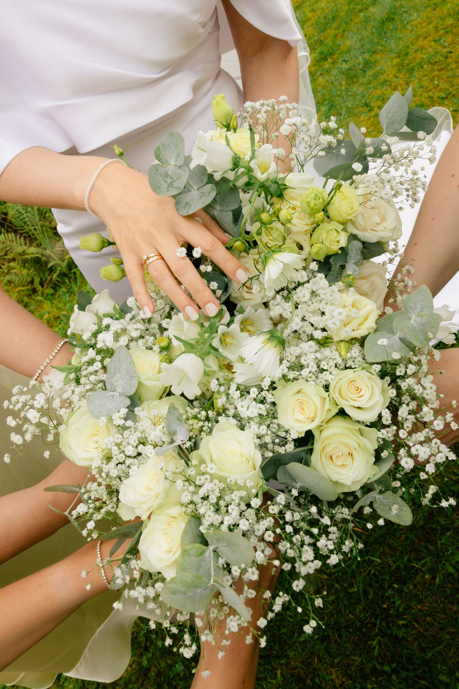 Bride holding elegant white rose and gypsophila wedding bouquet with eucalyptus, wearing a gold ring and bracelet.