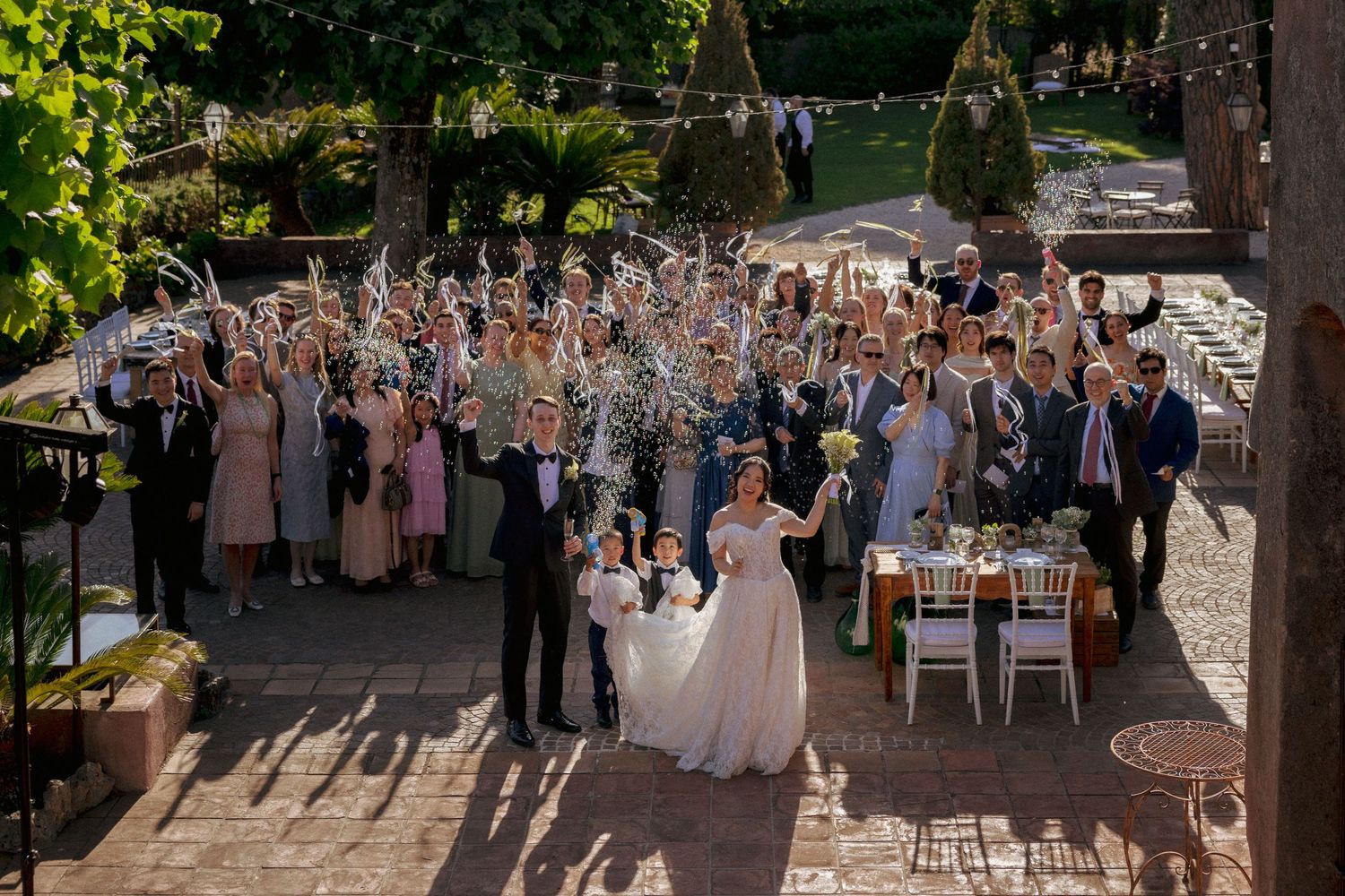 Bride and groom stand in a sunlit courtyard as wedding guests in formal attire cheer and toss flowers in celebration. Italia