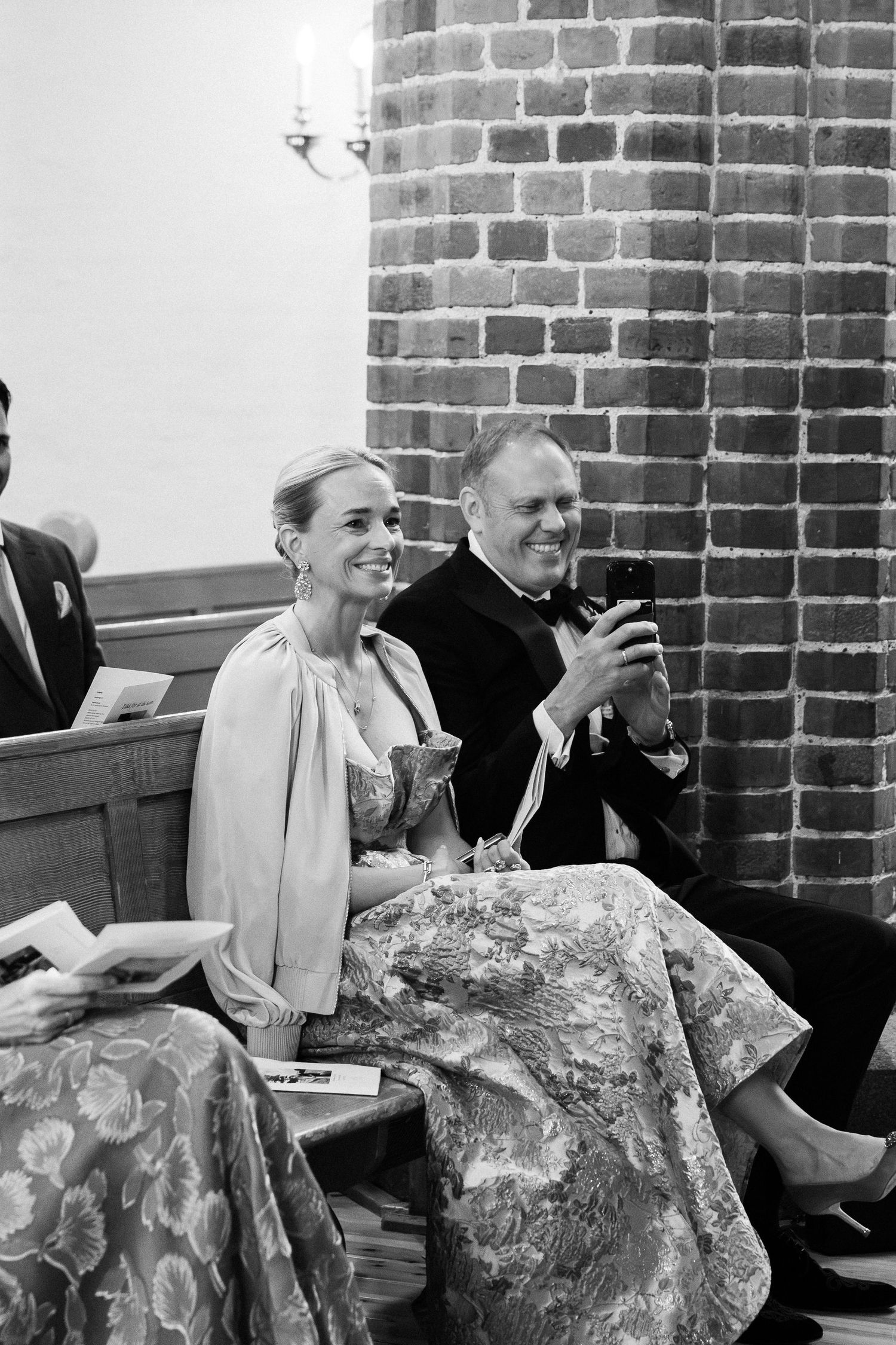 Black and white photo of smiling wedding guests seated in church pew, woman in floral dress beside man in tuxedo.