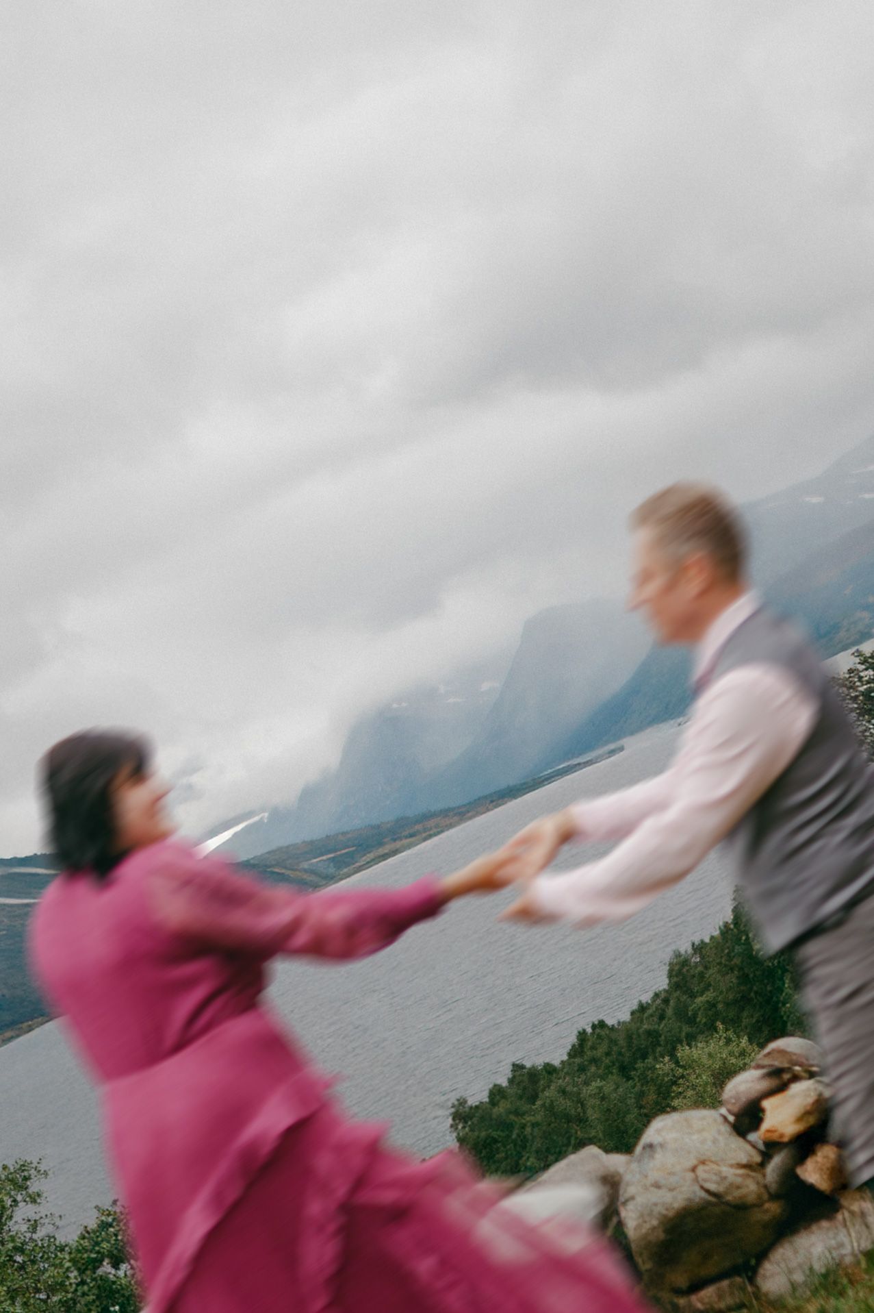 Couple dancing joyfully on a rocky overlook with misty fjord mountains in the background, woman in pink dress.