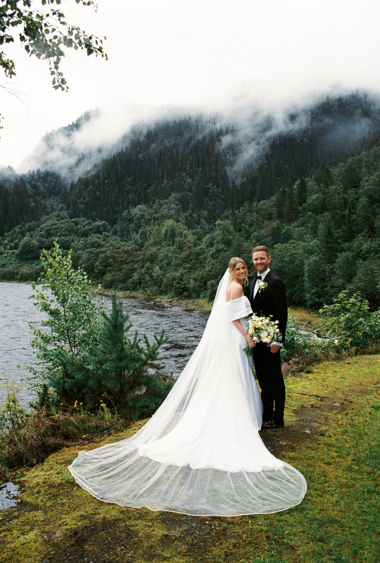 Bride and groom pose by misty mountain lake, her cathedral veil trailing on grass, holding white bouquet.