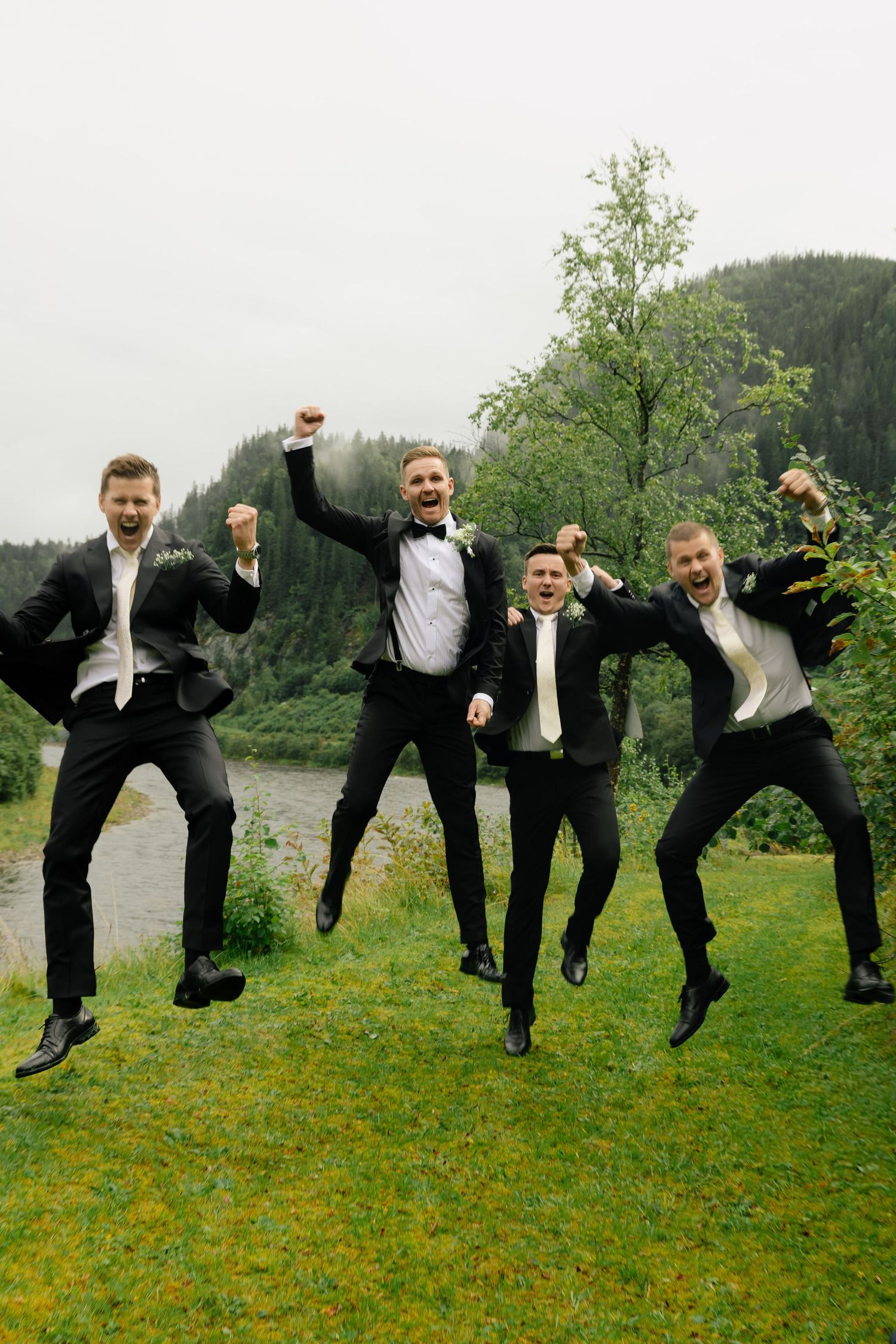 Four groomsmen in black tuxedos joyfully jumping outdoors with misty mountain and river backdrop.