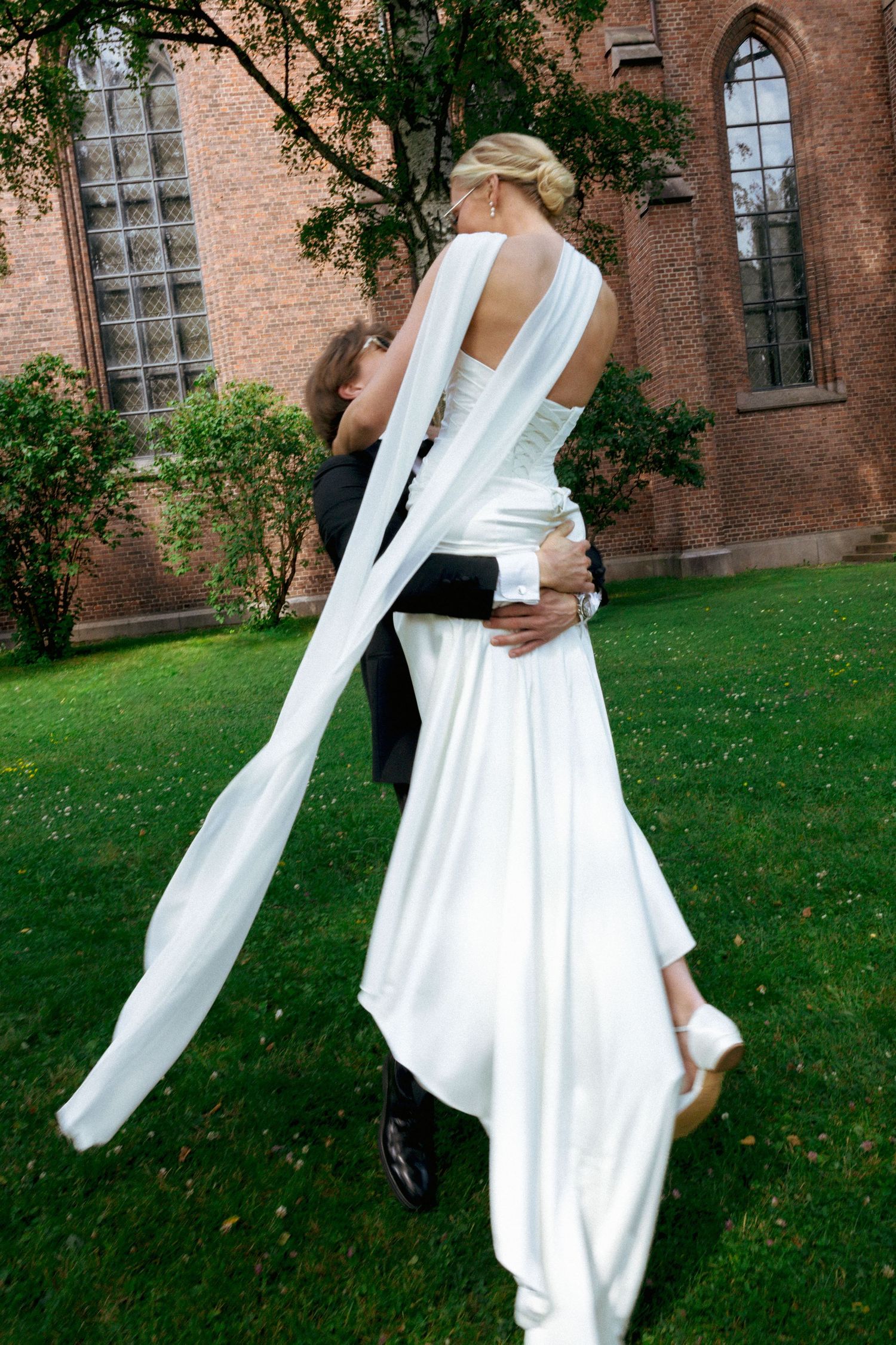 Groom lifting bride in white wedding gown and veil outside brick church on green lawn.