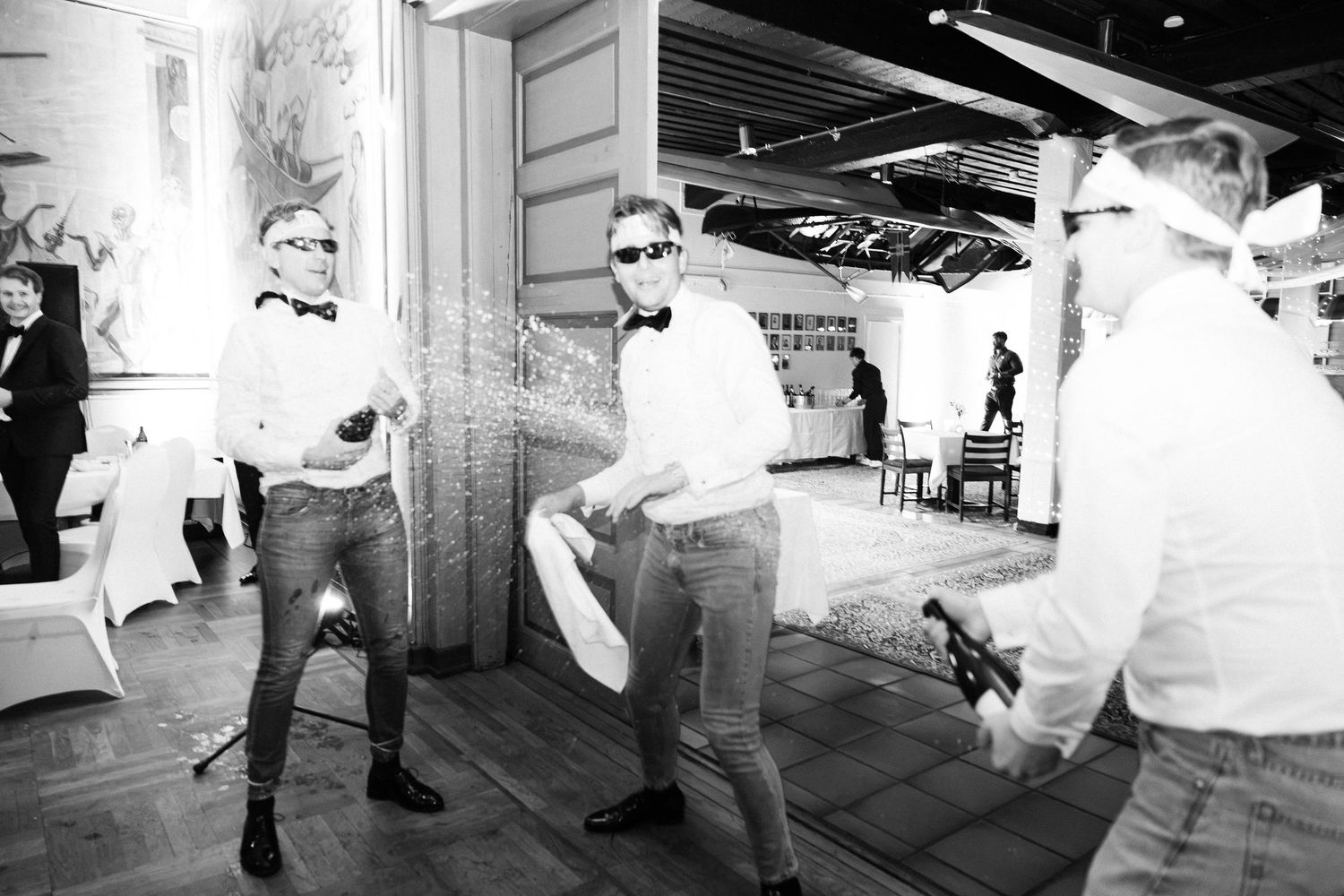 Black and white photo of men in bow ties spraying champagne bottles at wedding party.