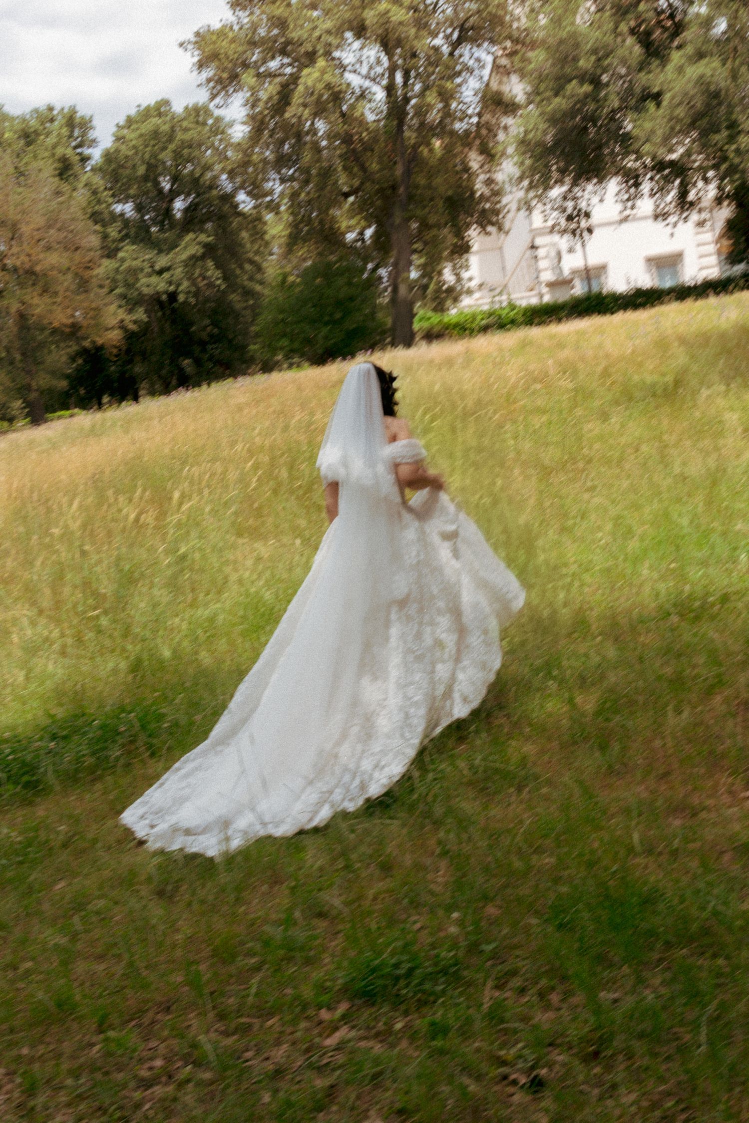 Bride in white lace wedding gown and veil walking through green meadow with trees and building in background.