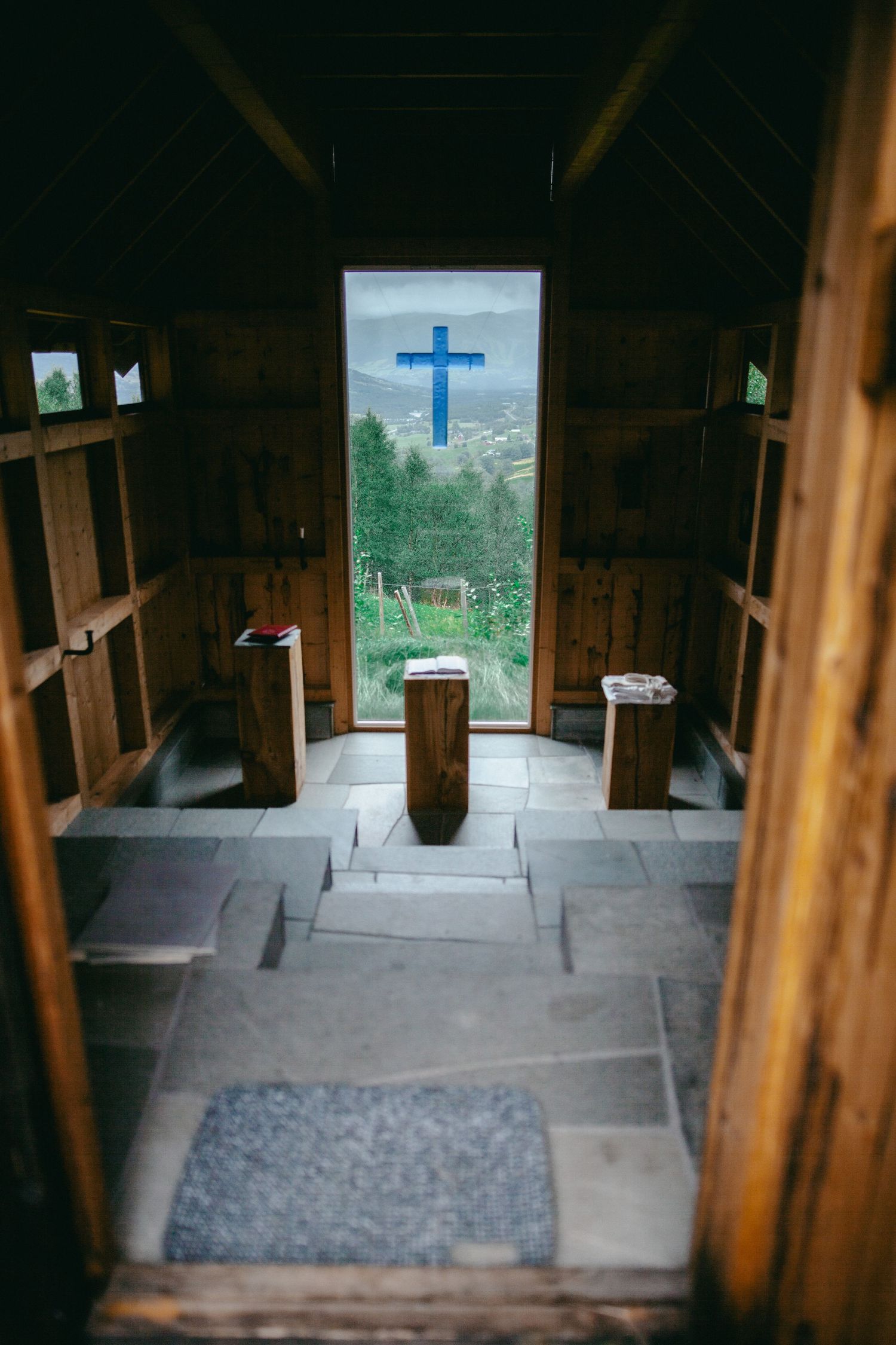 Interior of a rustic wooden chapel with stone floor, altar, and a blue cross visible through the open door with mountain views.