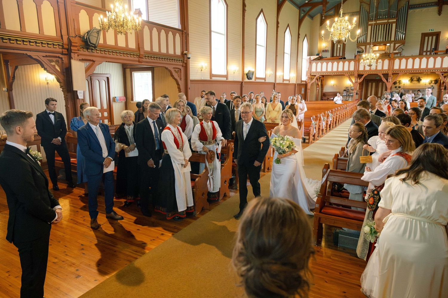 Guests stand in wooden church pews during an elegant wedding ceremony with chandeliers and stained glass windows.