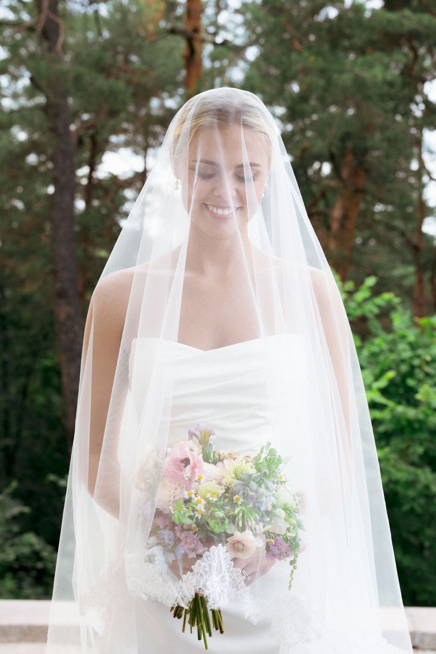 Smiling bride in white strapless gown holding floral bouquet under a flowing veil outdoors.