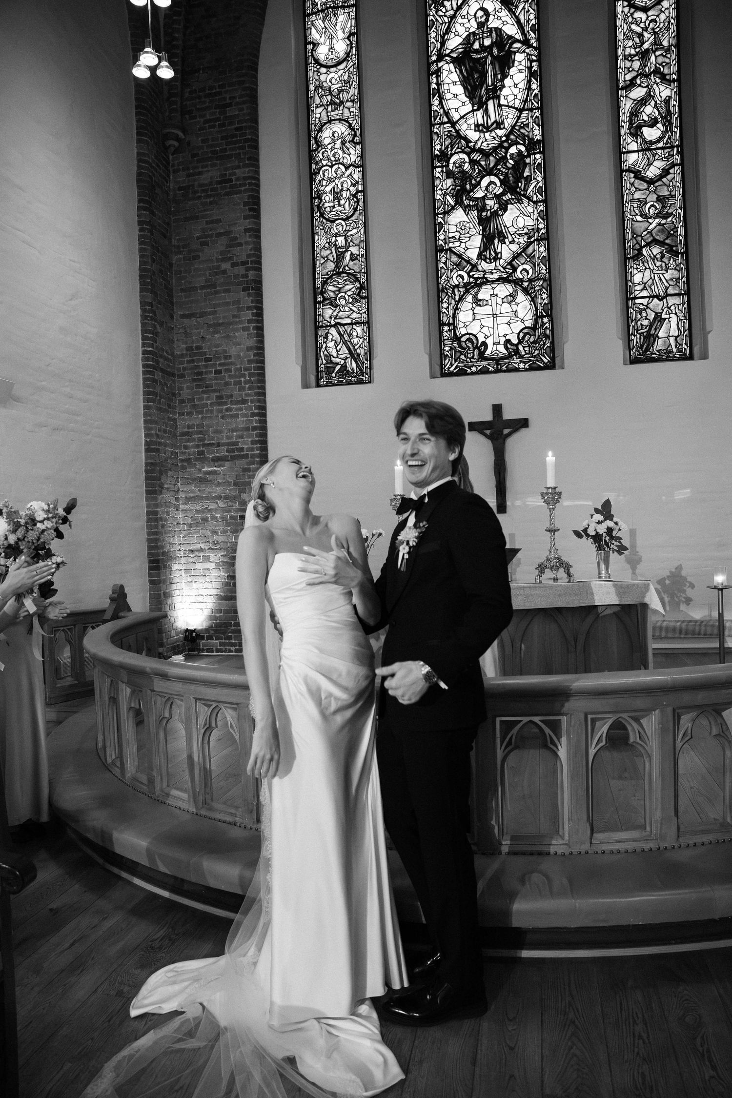 Black and white photo of bride and groom laughing at church altar with stained glass windows.