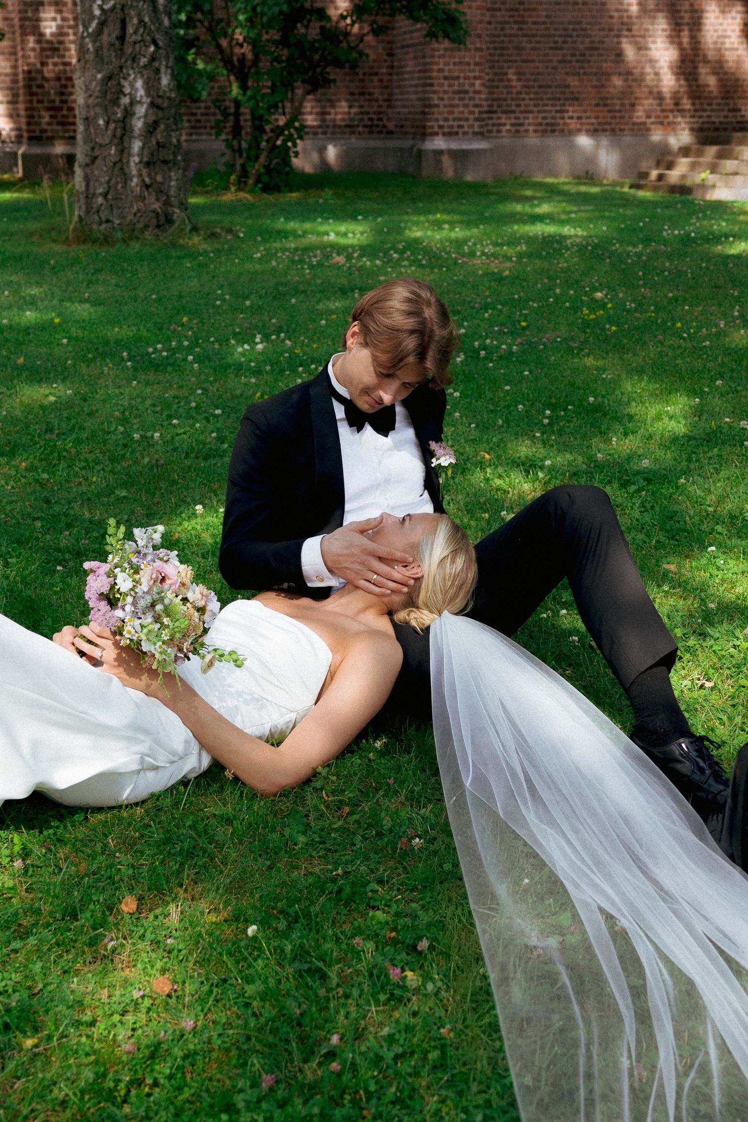 Bride in white gown lies on grass as groom in tuxedo tenderly holds her face during outdoor wedding photo session.