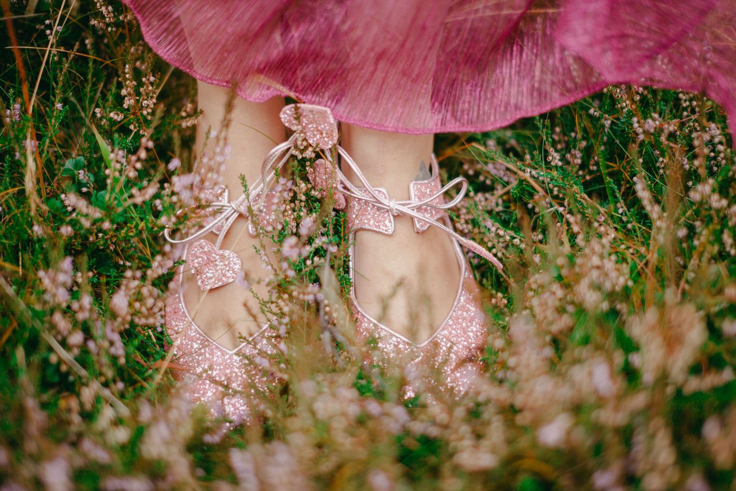 Close-up of sparkling pink glitter ballet flats with bow ties on a woman's feet in a lush green meadow.