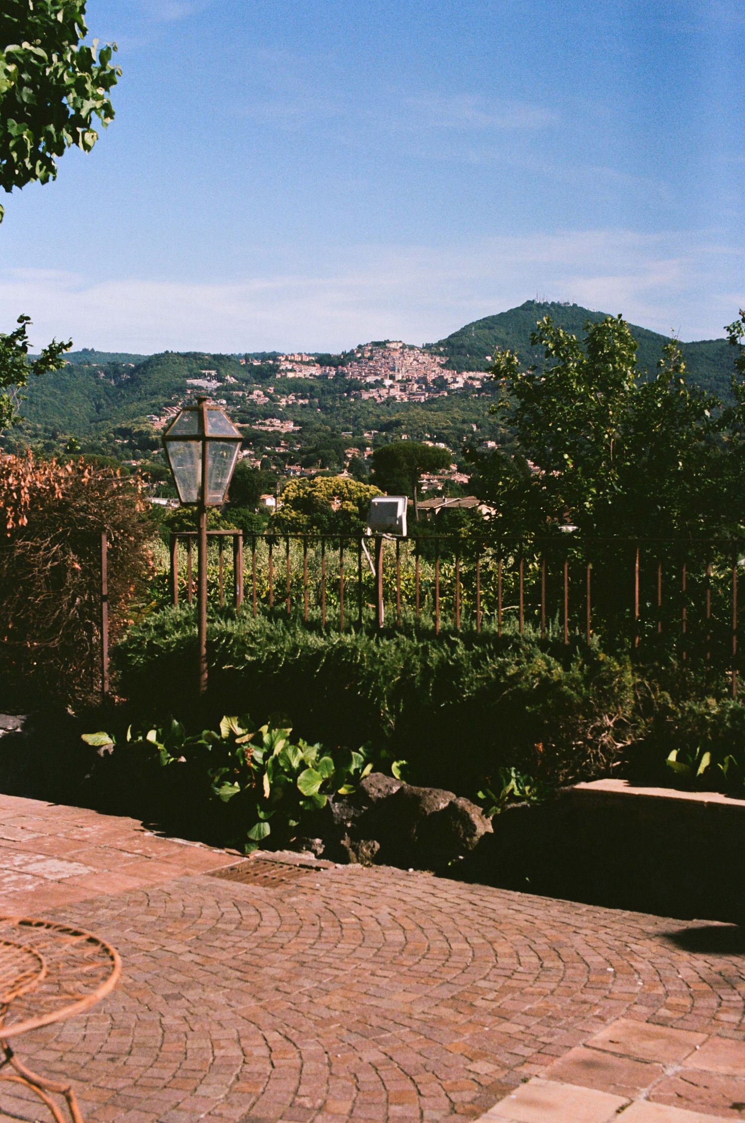Scenic Italian hillside garden with cobblestone terrace, lush greenery, and a historic village nestled among rolling mountains.