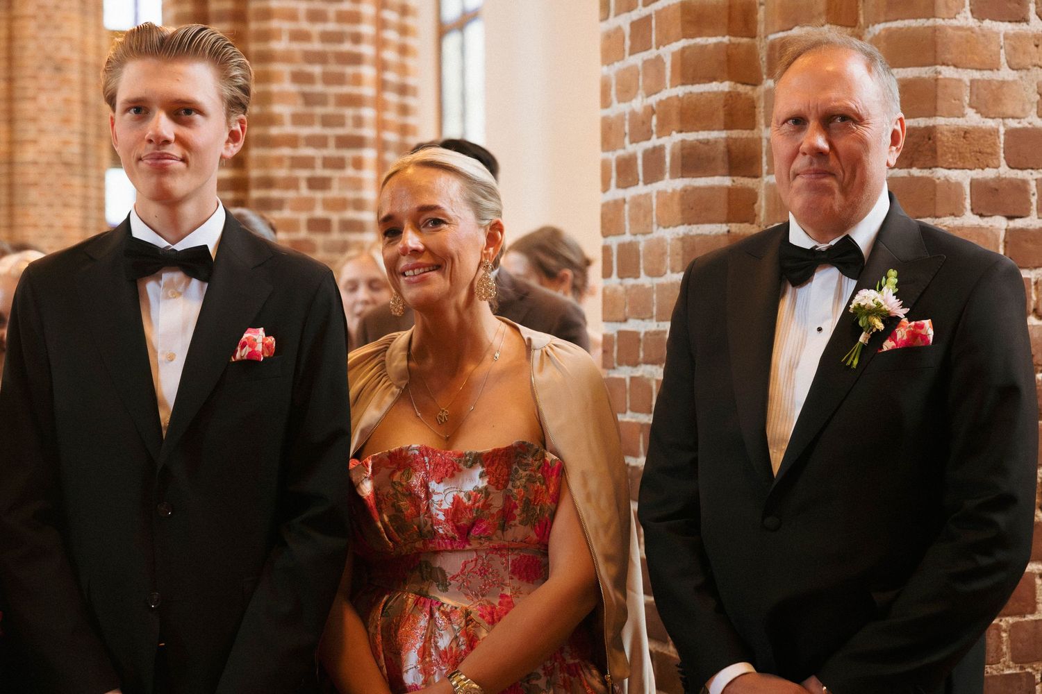 Three elegantly dressed people in formal attire pose together at a wedding ceremony in a brick-walled venue.