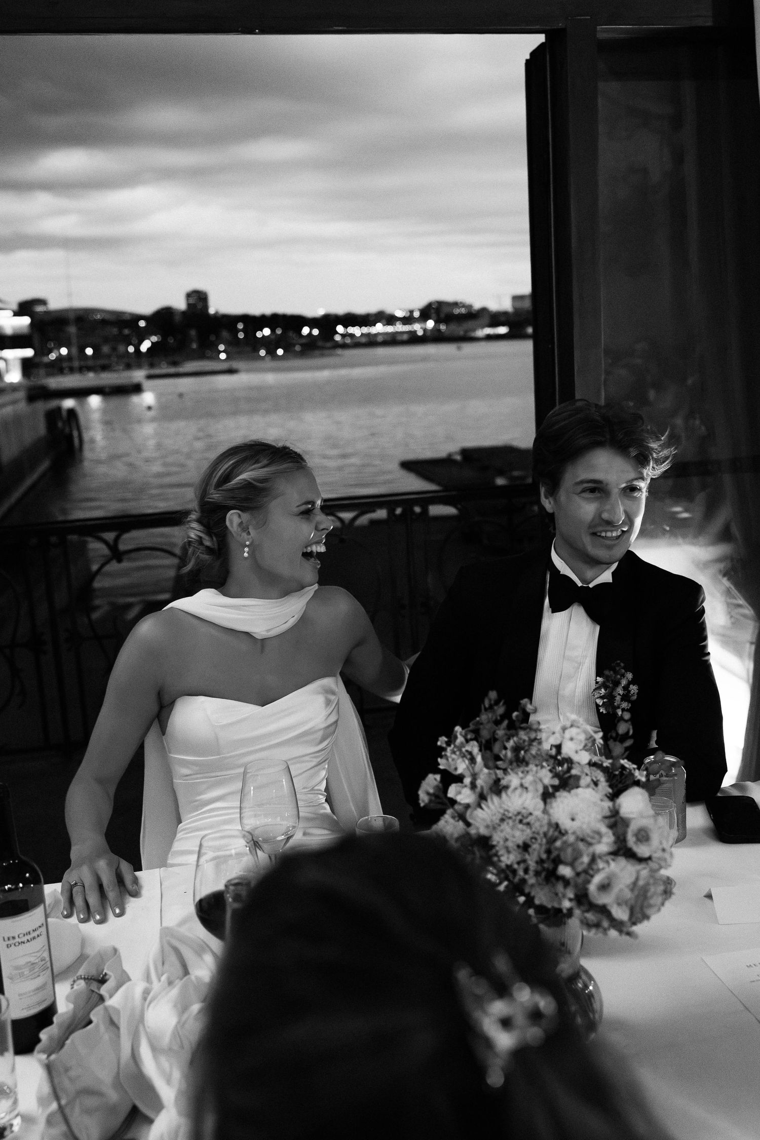 Bride and groom laughing at wedding reception table with waterfront view at dusk.