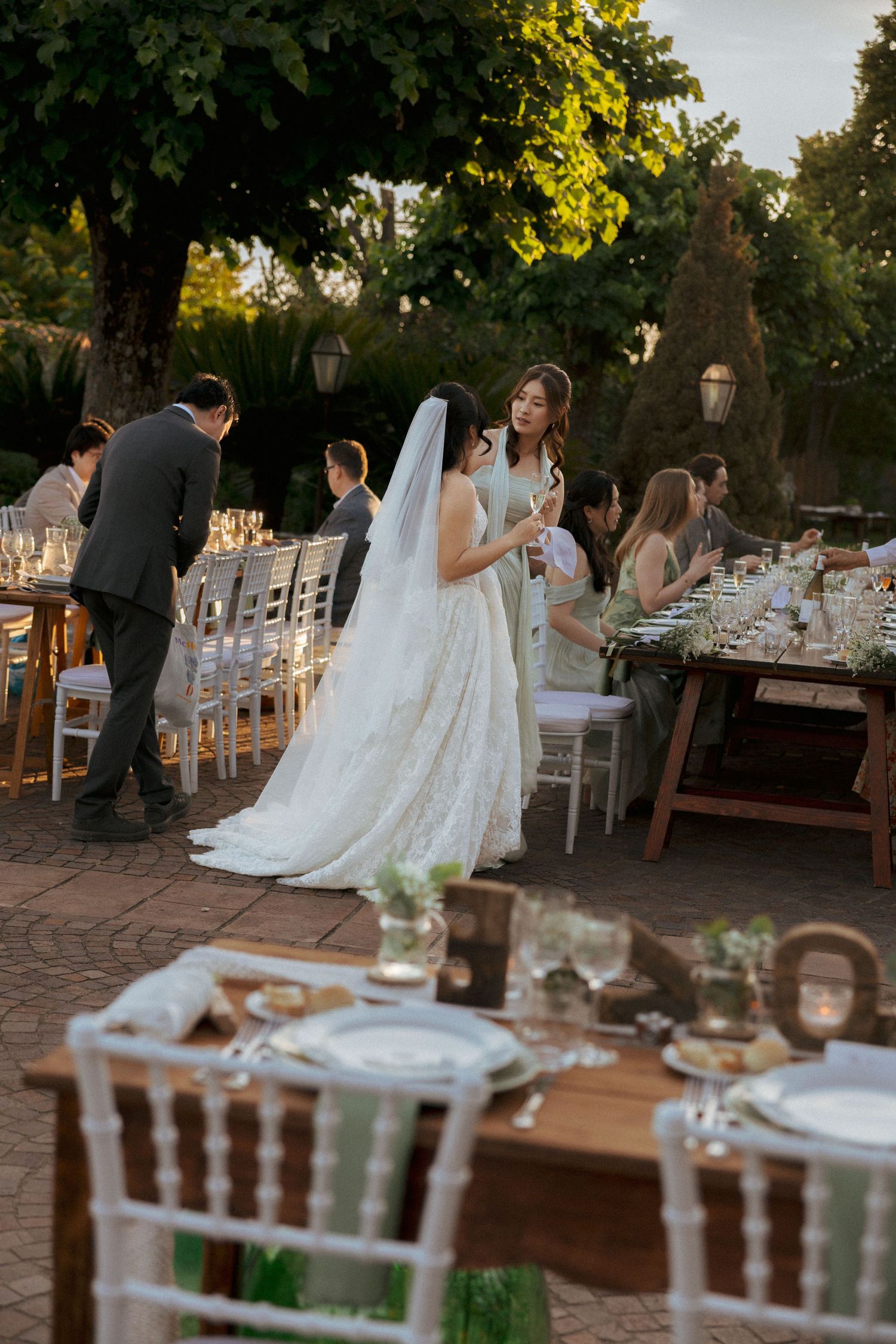 Bride in white lace gown and veil mingles with guests at elegant outdoor wedding reception.