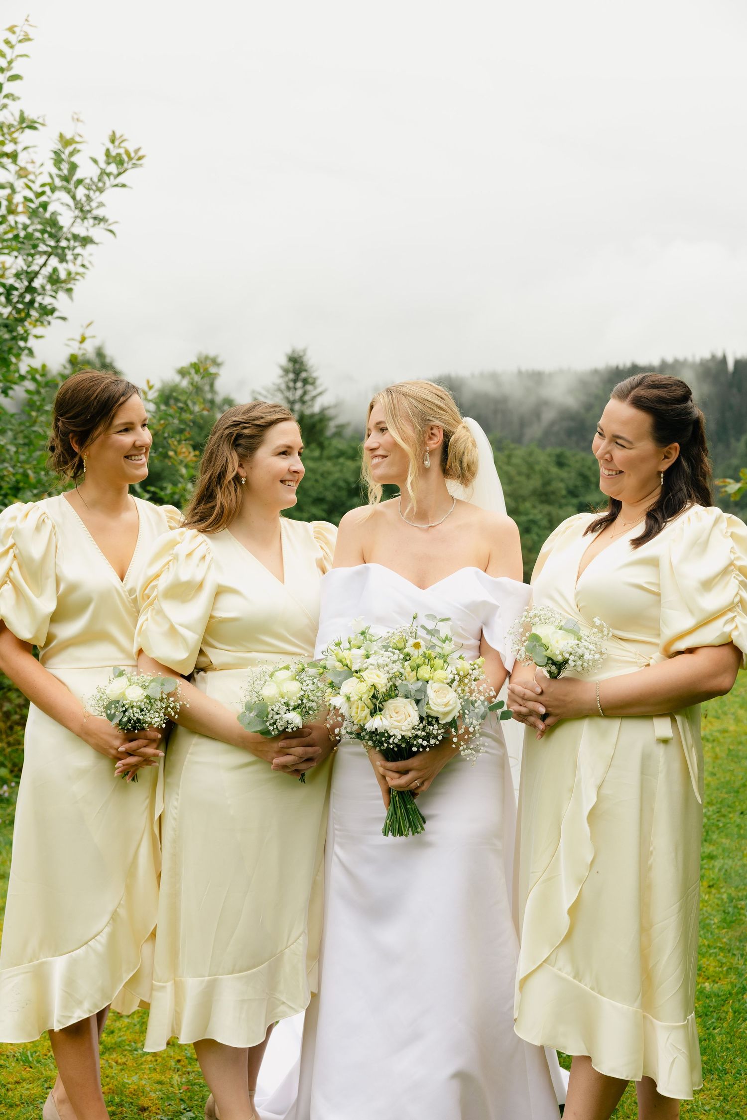 Bride in white strapless gown with three bridesmaids in yellow dresses holding white bouquets outdoors.