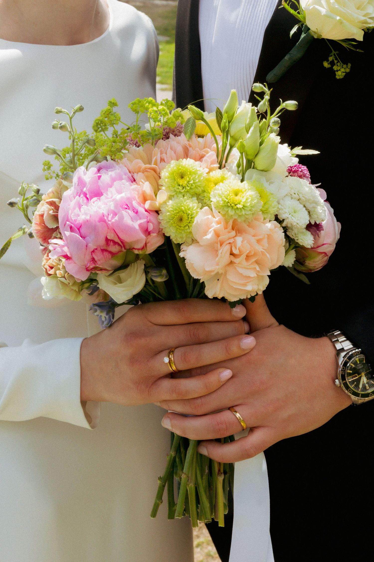 Bride and groom holding a colorful wedding bouquet with pink peonies, peach roses, and green flowers, both wearing gold rings.