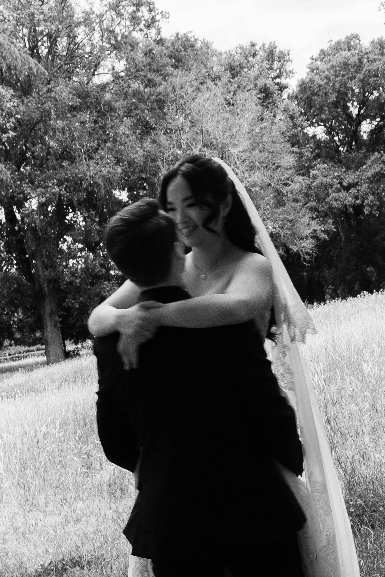 Black and white photo of a bride in veil embracing groom outdoors among trees on their wedding day.