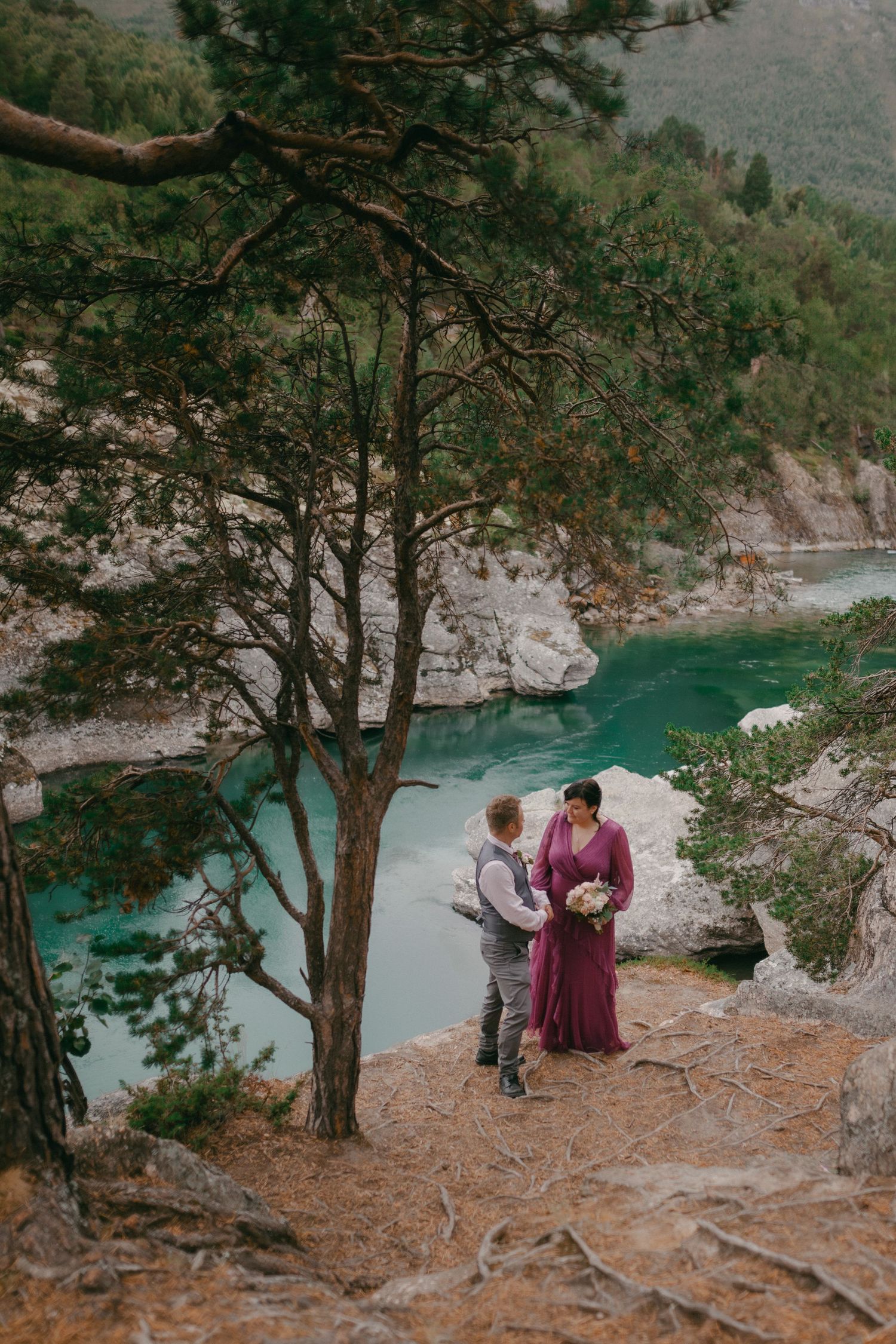 Bride and groom standing beneath pine tree overlooking stunning turquoise river canyon during mountain elopement.
