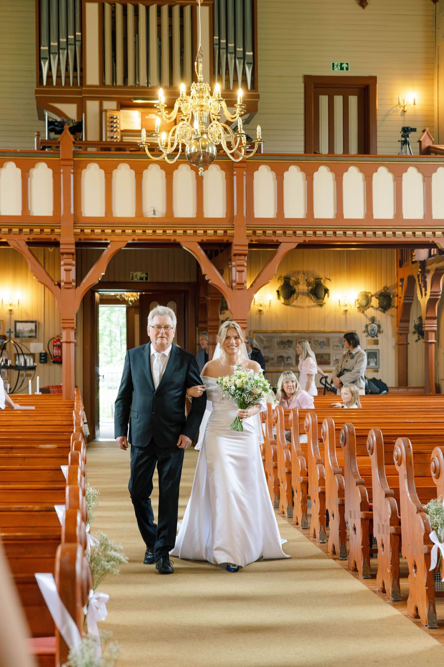 A bride walks down the church aisle with her father during a wedding ceremony in a wooden chapel.