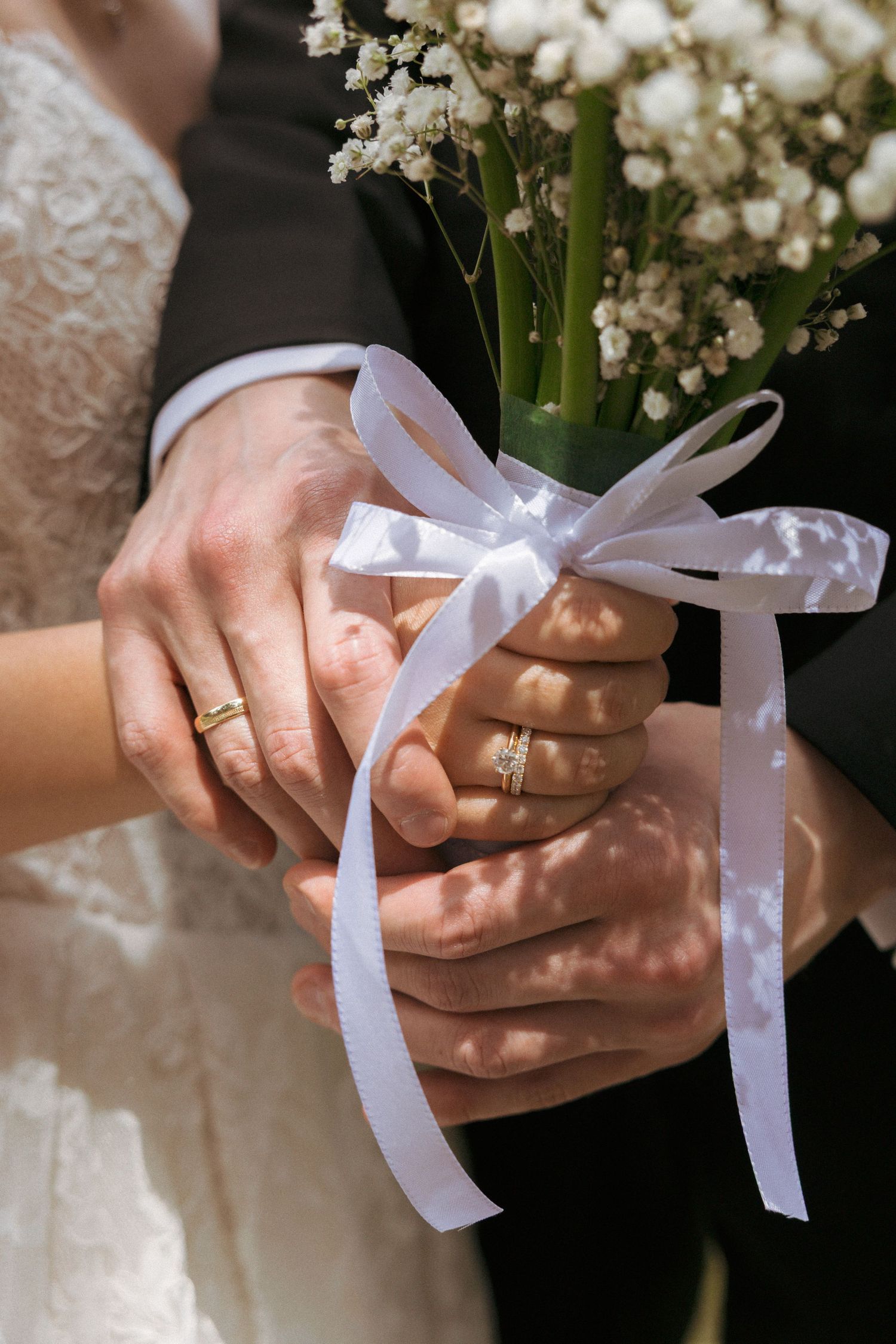 Newlywed couple holding a white ribbon-tied bouquet of baby's breath, showing gold wedding rings on their hands.