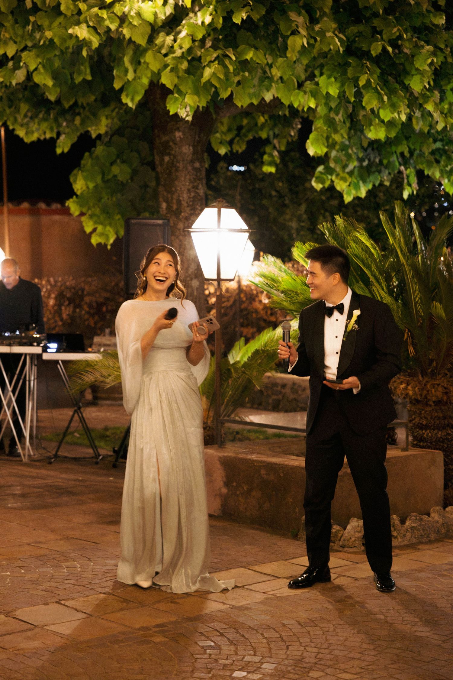 Bride in white gown and groom in tuxedo laughing together outdoors at evening wedding reception.