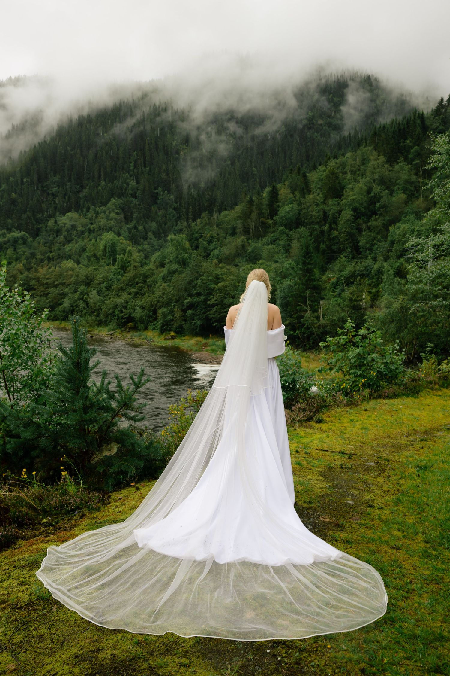 Bride in white gown and long veil stands before misty mountain forest and river landscape.