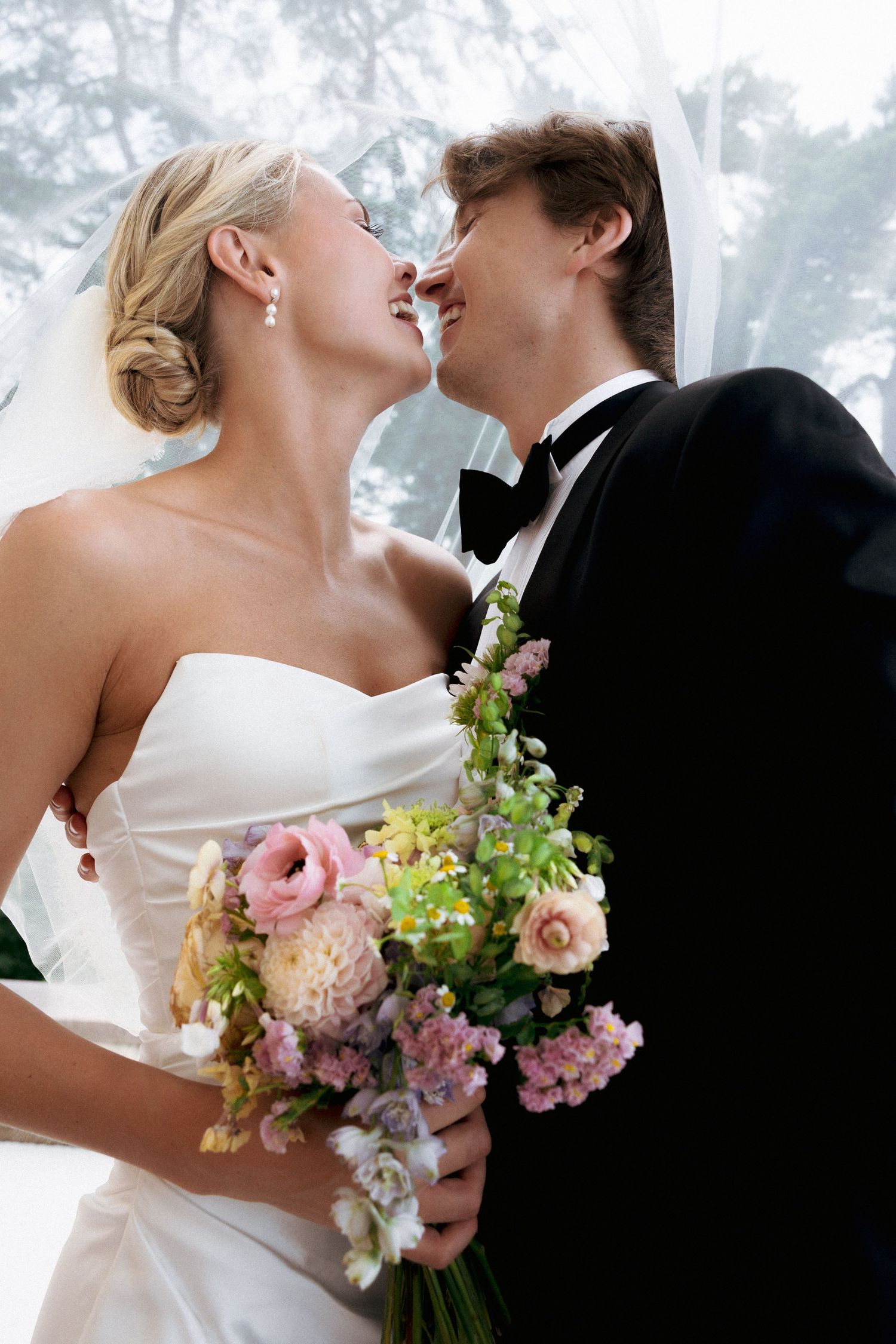 A joyful bride and groom share a romantic moment, laughing together while holding a pastel wedding bouquet.