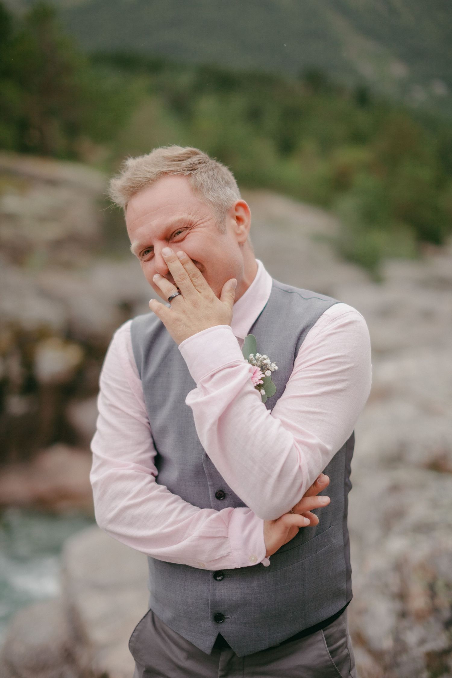 Laughing groom in gray vest and pink shirt with boutonniere covering his smile during wedding celebration outdoors.